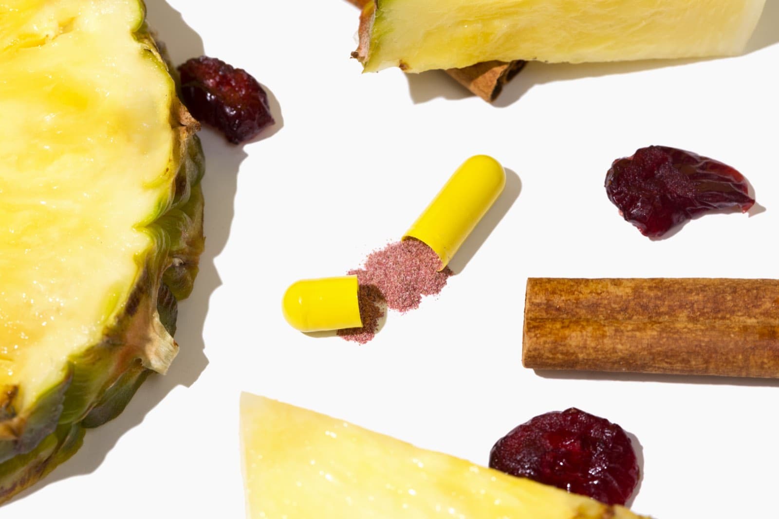 An open yellow capsule beside pineapple, cranberries, and cinnamon on white