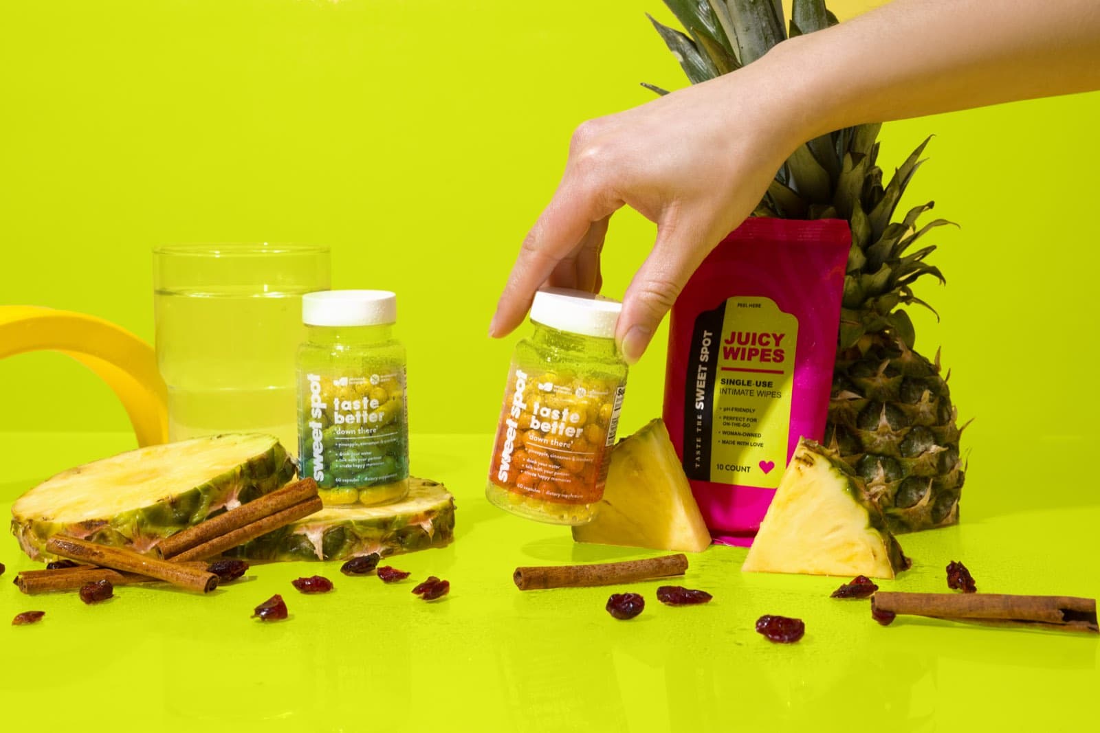 A hand lifts the For Her bottle from a lineup with For Him, wipes, pineapple, and water