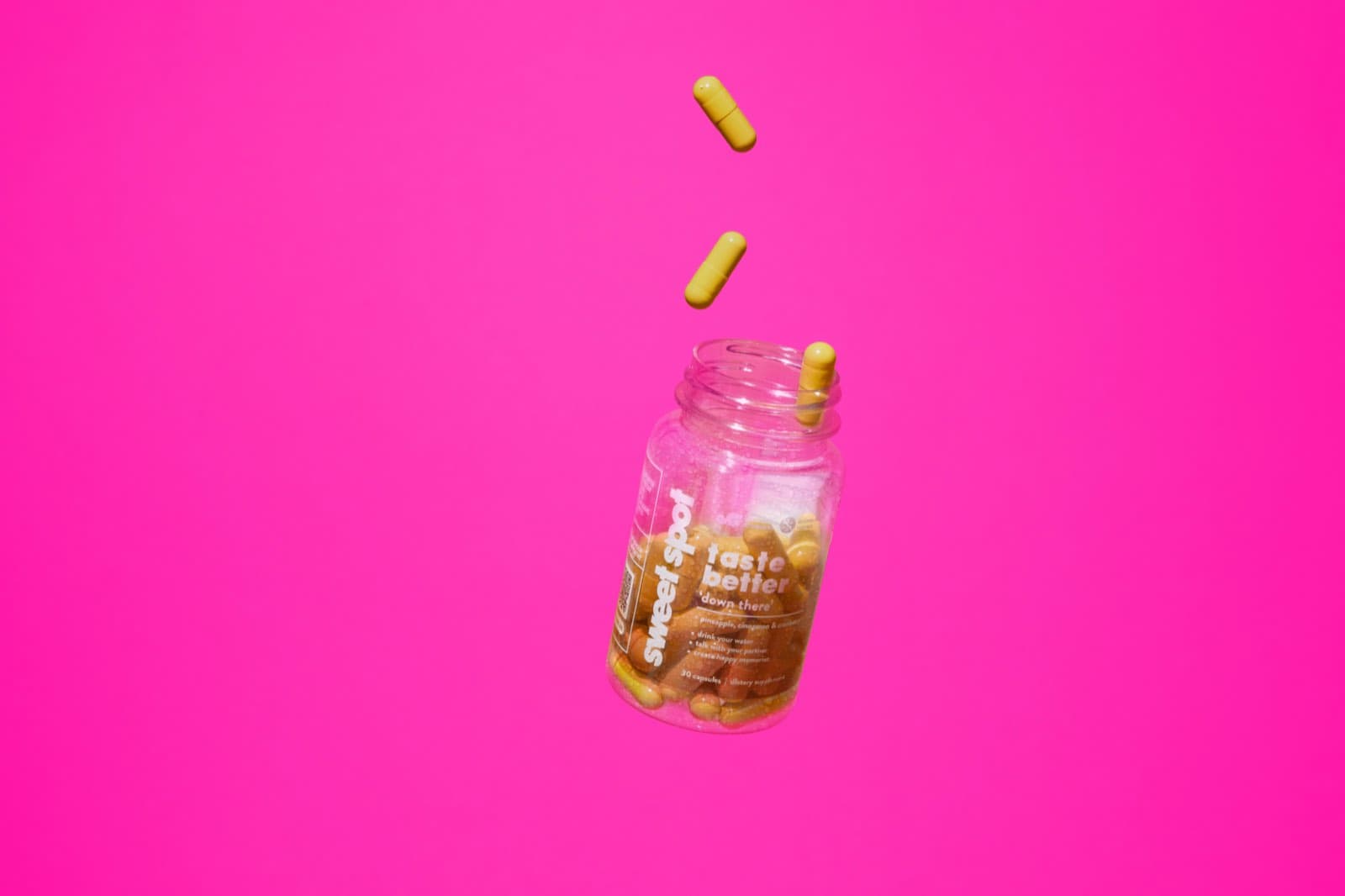 Yellow capsules falling toward an open For Her bottle against pink