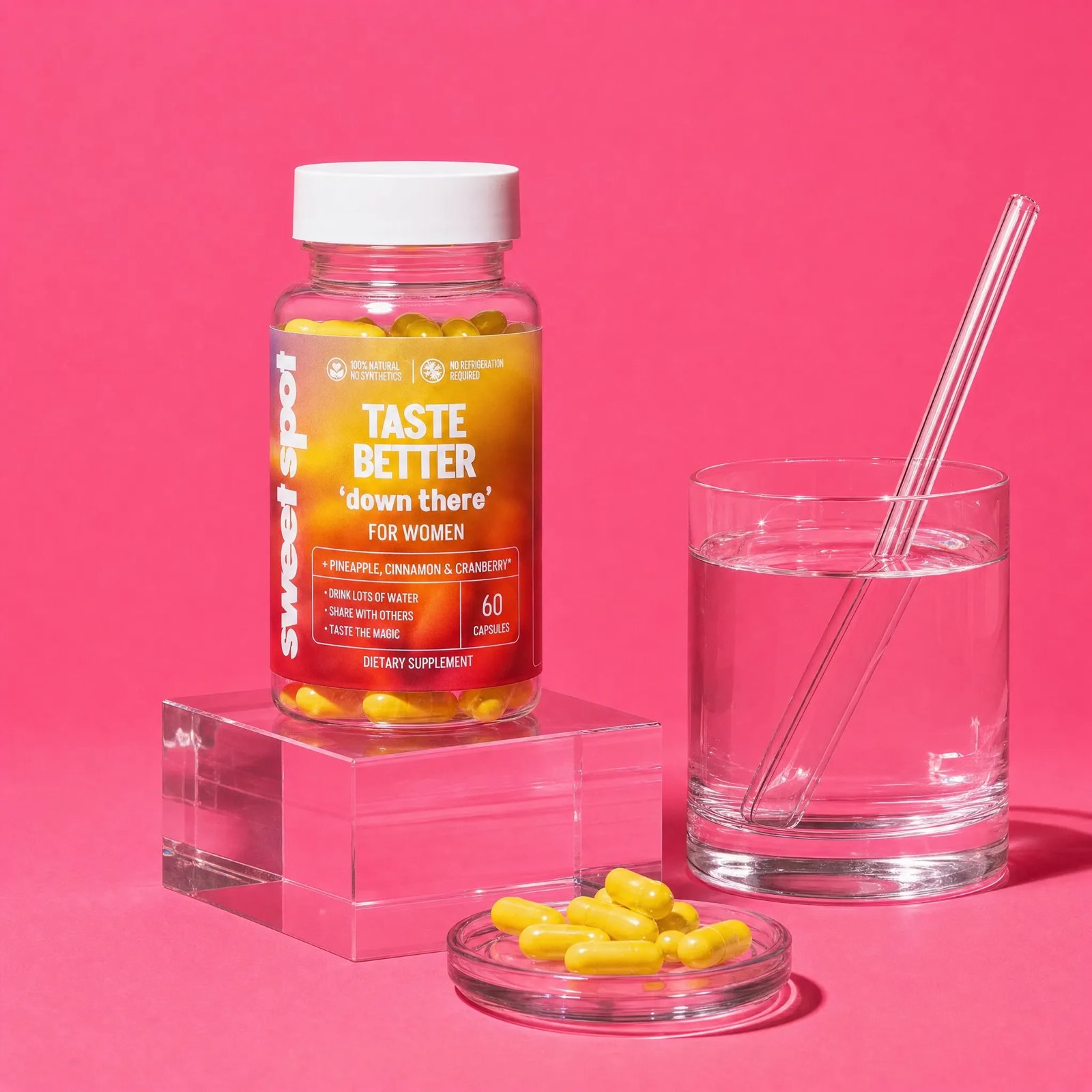 A Sweet Spot bottle labeled taste better down there for women sits on a clear block beside a water glass and yellow capsules on a pink set.