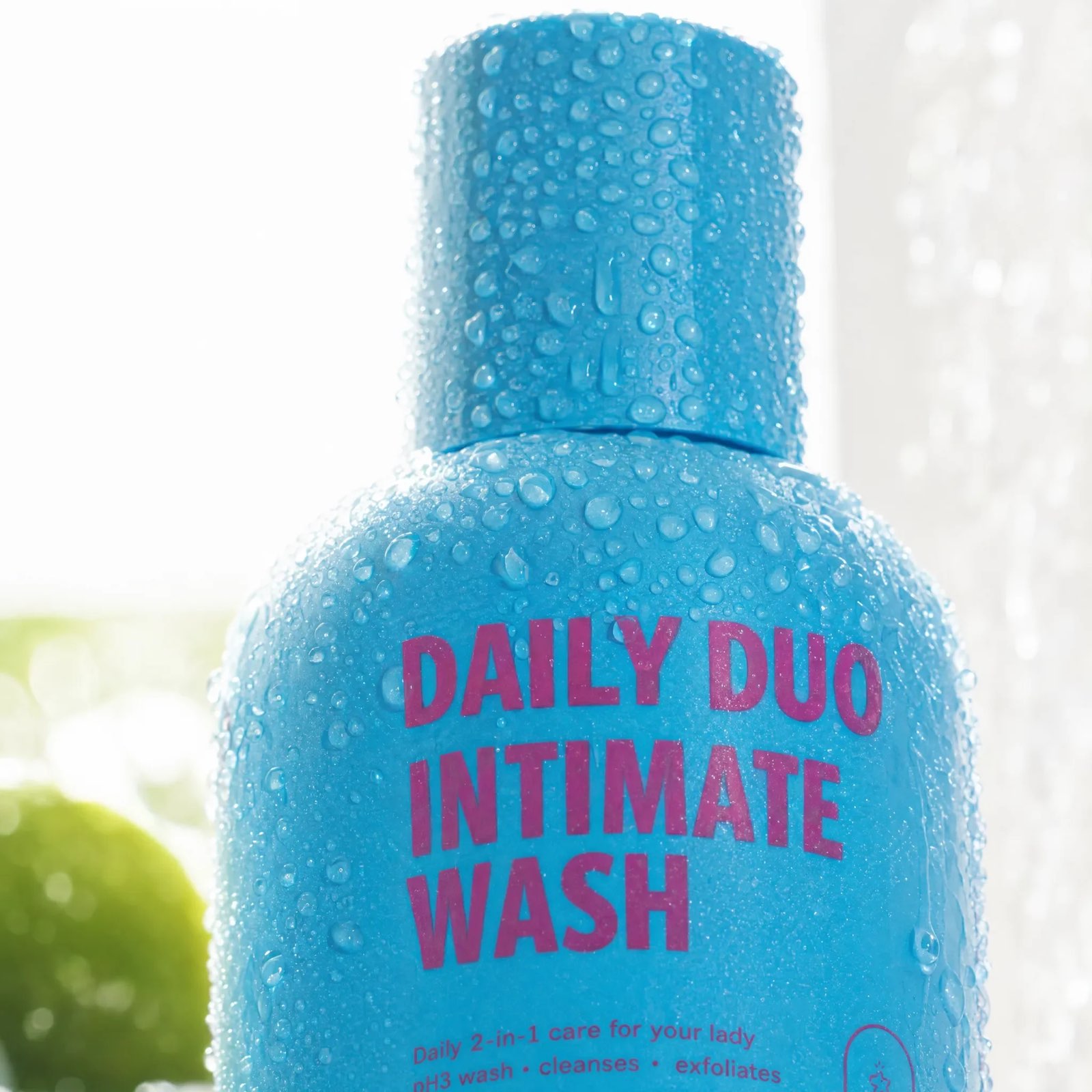 Water beads cover a close view of a blue bottle whose label reads Daily Duo Intimate Wash against a bright wet window.