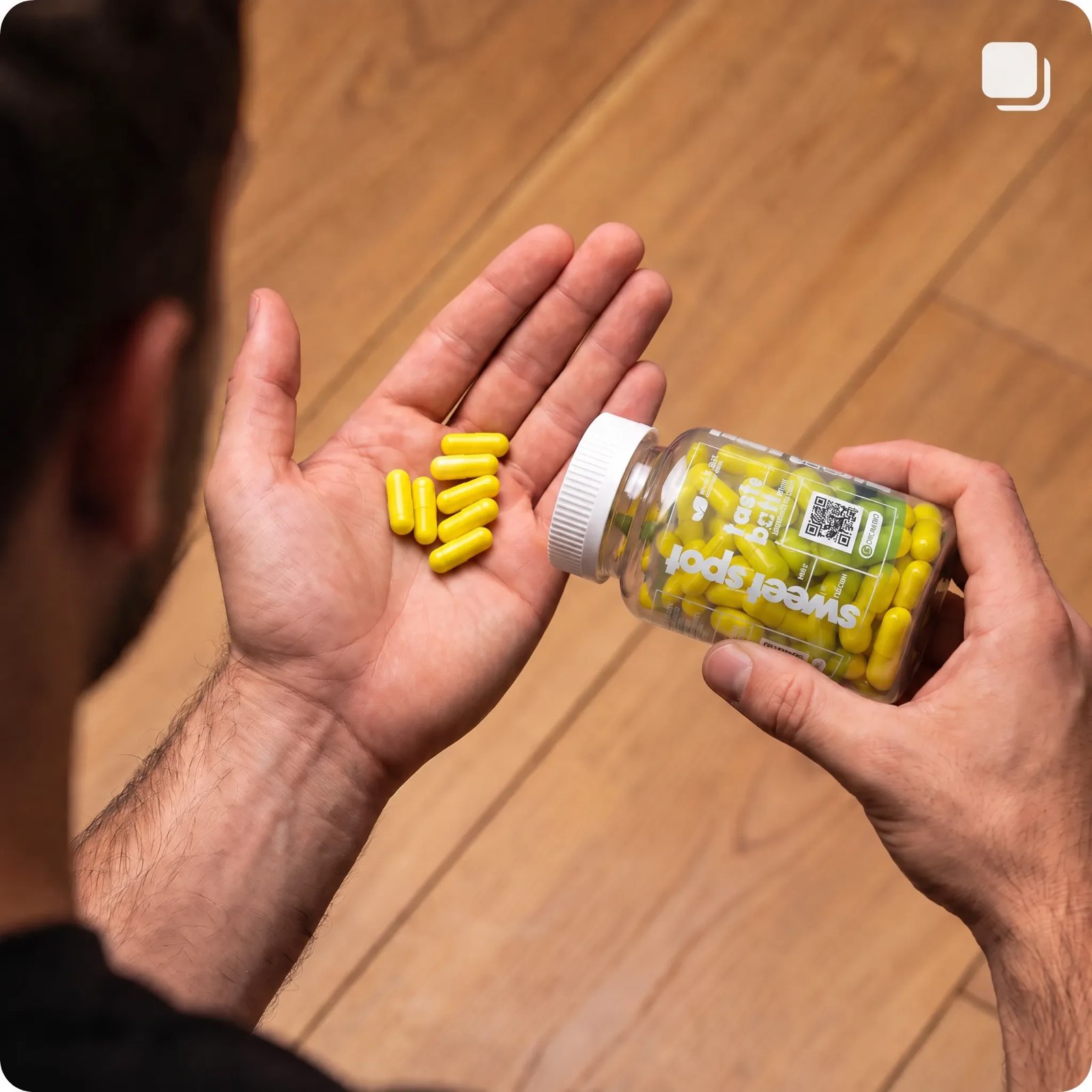 A man tips a Sweet Spot bottle of yellow capsules into his open palm over a wood floor, with the white cap still on.