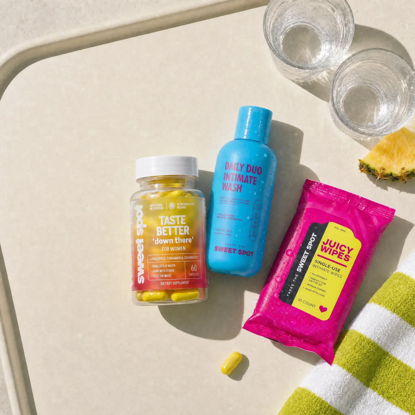 A cream tray holds a Taste Better down there For Women bottle, a blue Daily Duo Intimate Wash, pink Juicy Wipes, two water glasses, and a pineapple wedge.