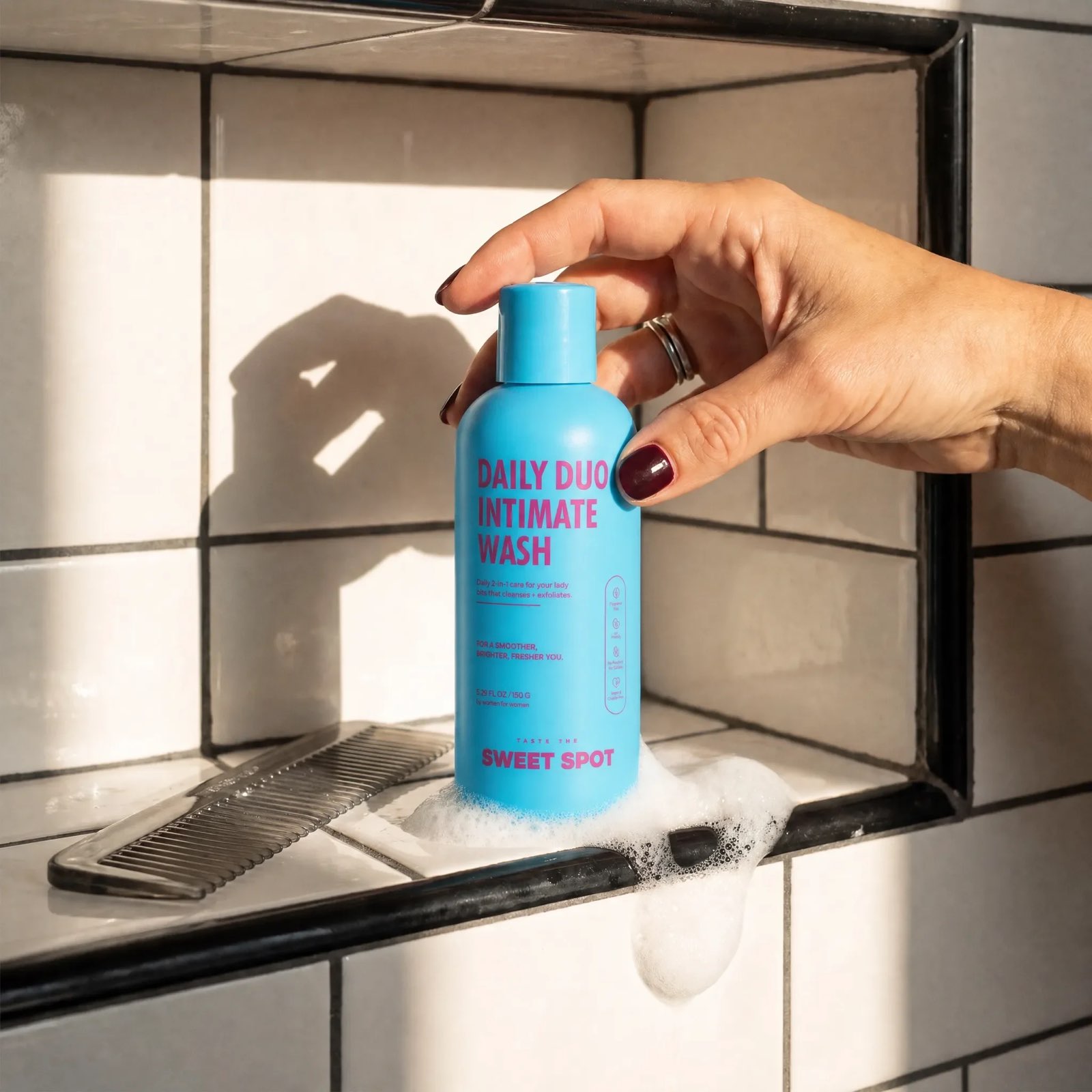 A hand with dark red nails sets a blue Daily Duo Intimate Wash bottle into soap foam on a white tiled ledge beside a black comb.
