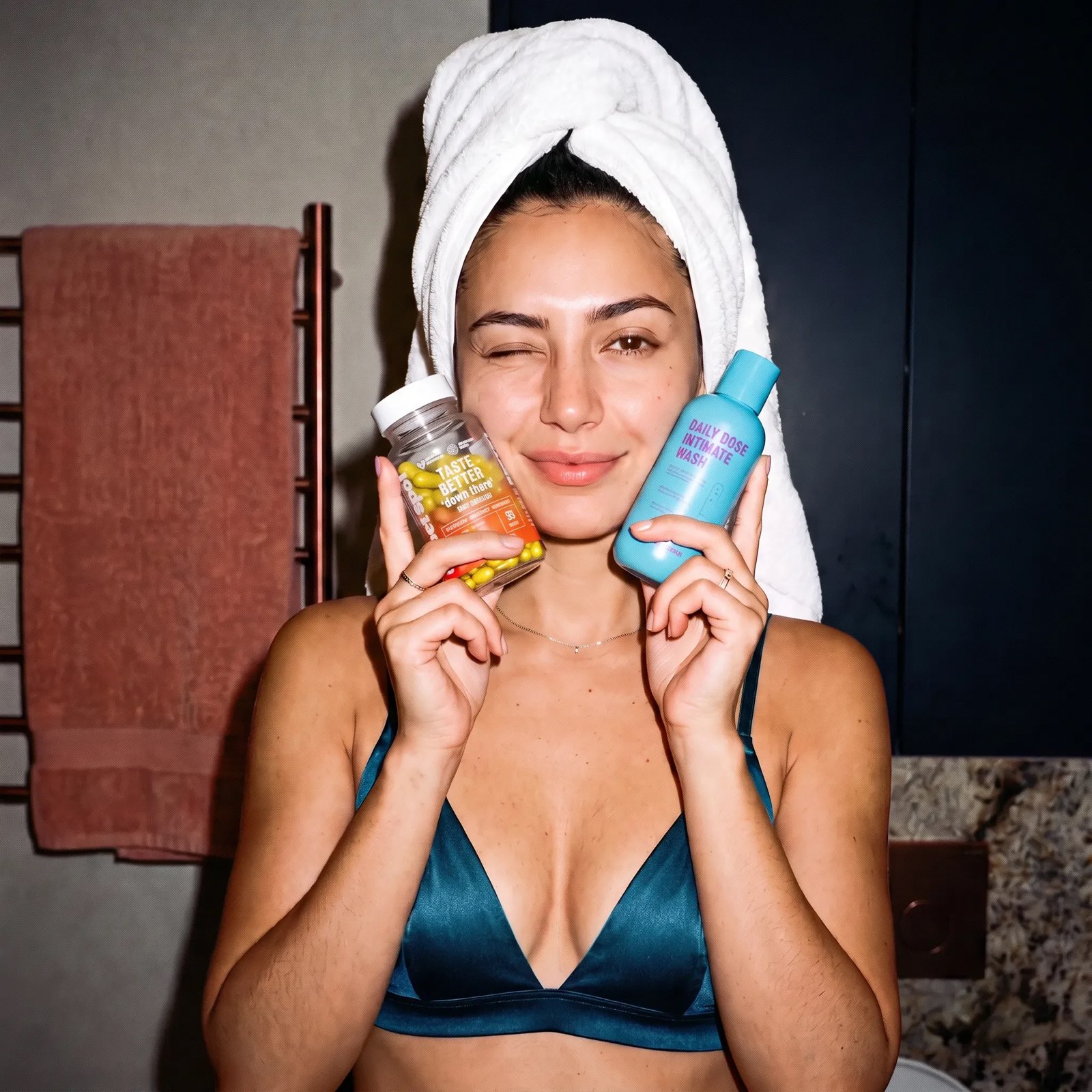 A woman in a teal top, towel on her head, winks while holding a Taste Better down there bottle and a blue Daily Dose Intimate Wash.