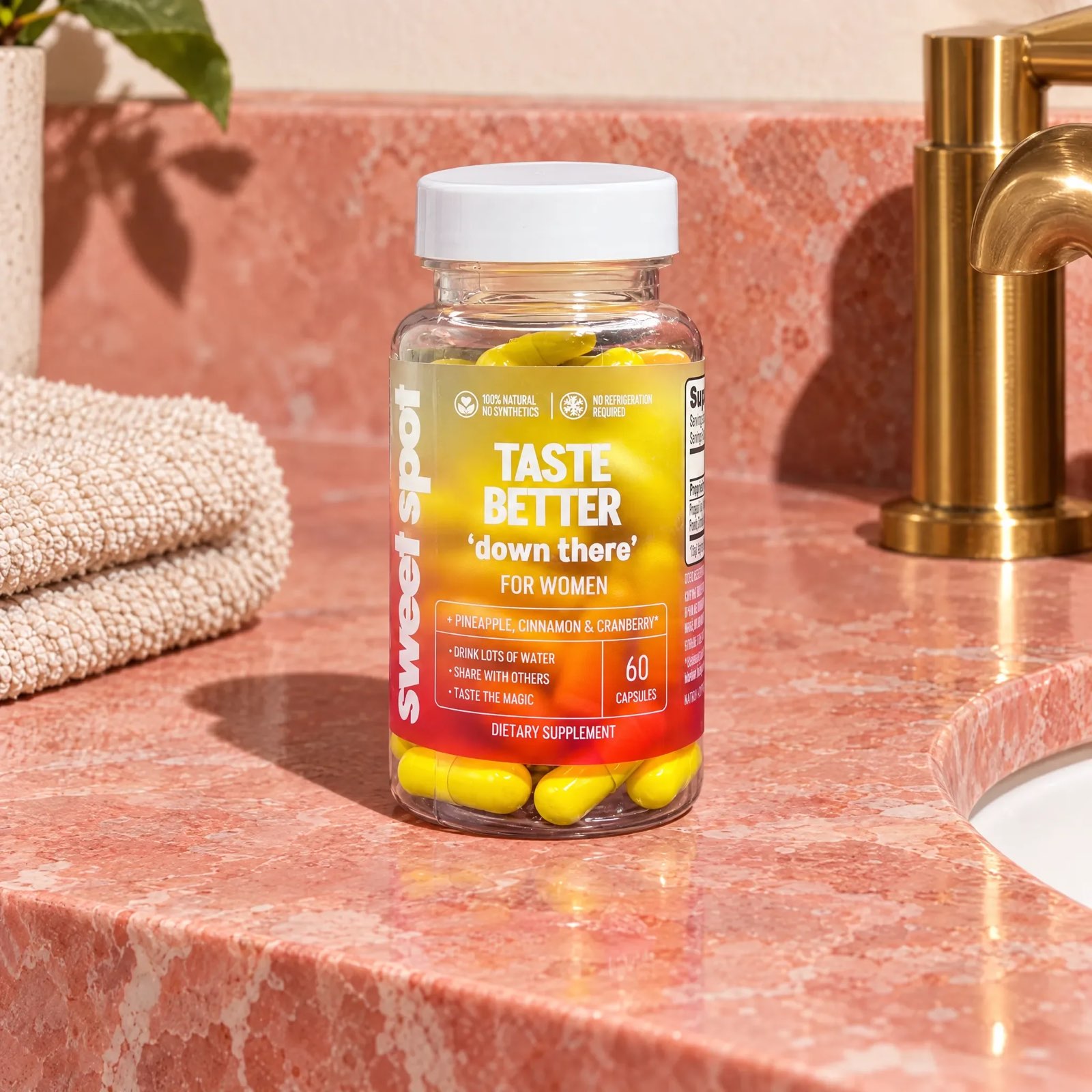 A Taste Better down there For Women bottle stands on a pink marble counter beside a gold faucet and a folded beige towel.