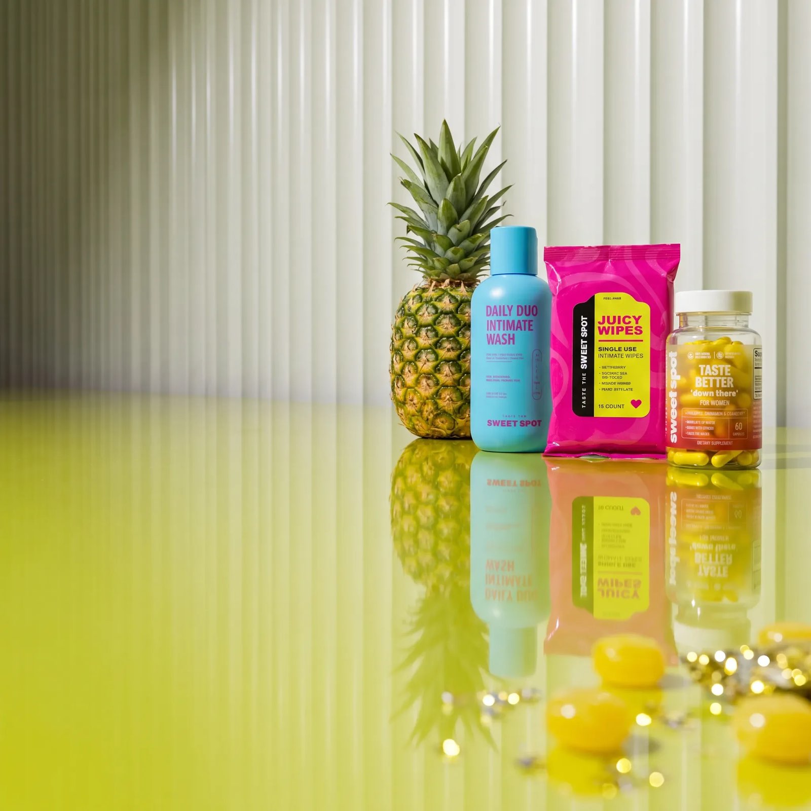 A pineapple, a blue Daily Duo Intimate Wash, pink Juicy Wipes, and a Taste Better down there For Women bottle stand on a glossy yellow surface.
