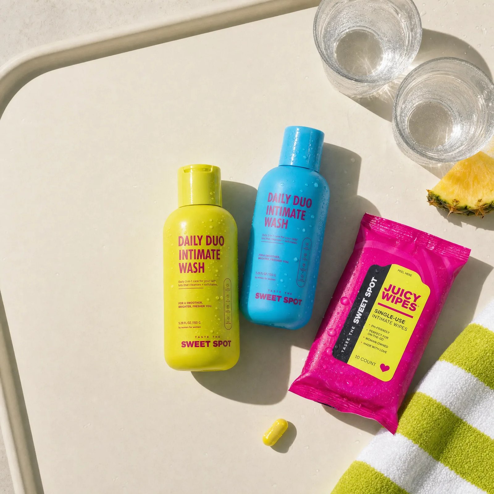 A cream tray holds a yellow Daily Duo Intimate Wash, a blue Daily Duo Intimate Wash, pink Juicy Wipes, two water glasses, and a pineapple wedge.