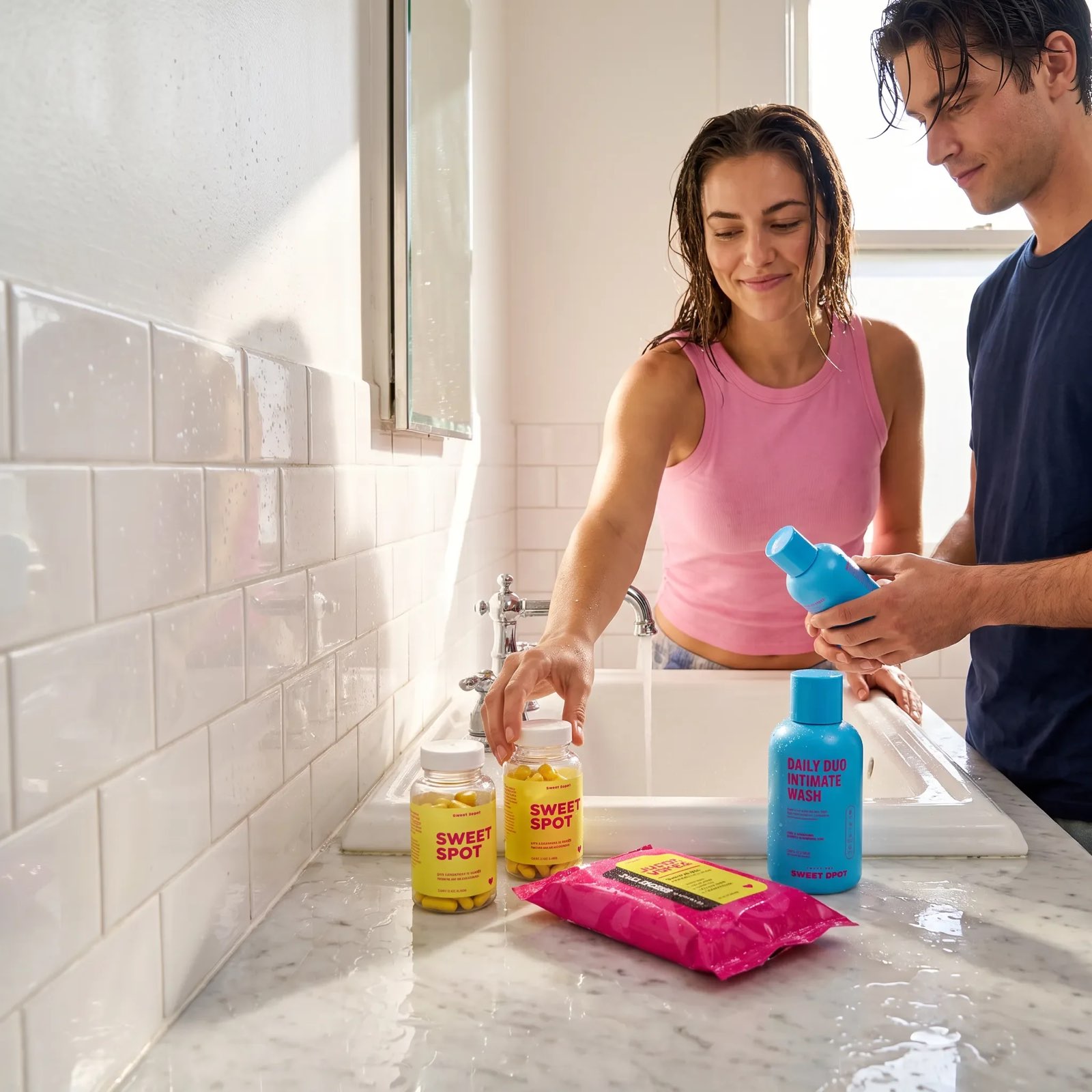 A woman in pink and a man in navy stand at a bathroom sink with Sweet Spot capsule bottles, pink wipes, and a blue Daily Duo wash.