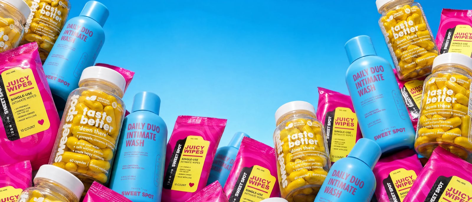 Yellow Sweet Spot taste better down there bottles, blue Daily Duo Intimate Wash bottles, and pink Juicy Wipes packets crowd both edges of a clear blue sky.