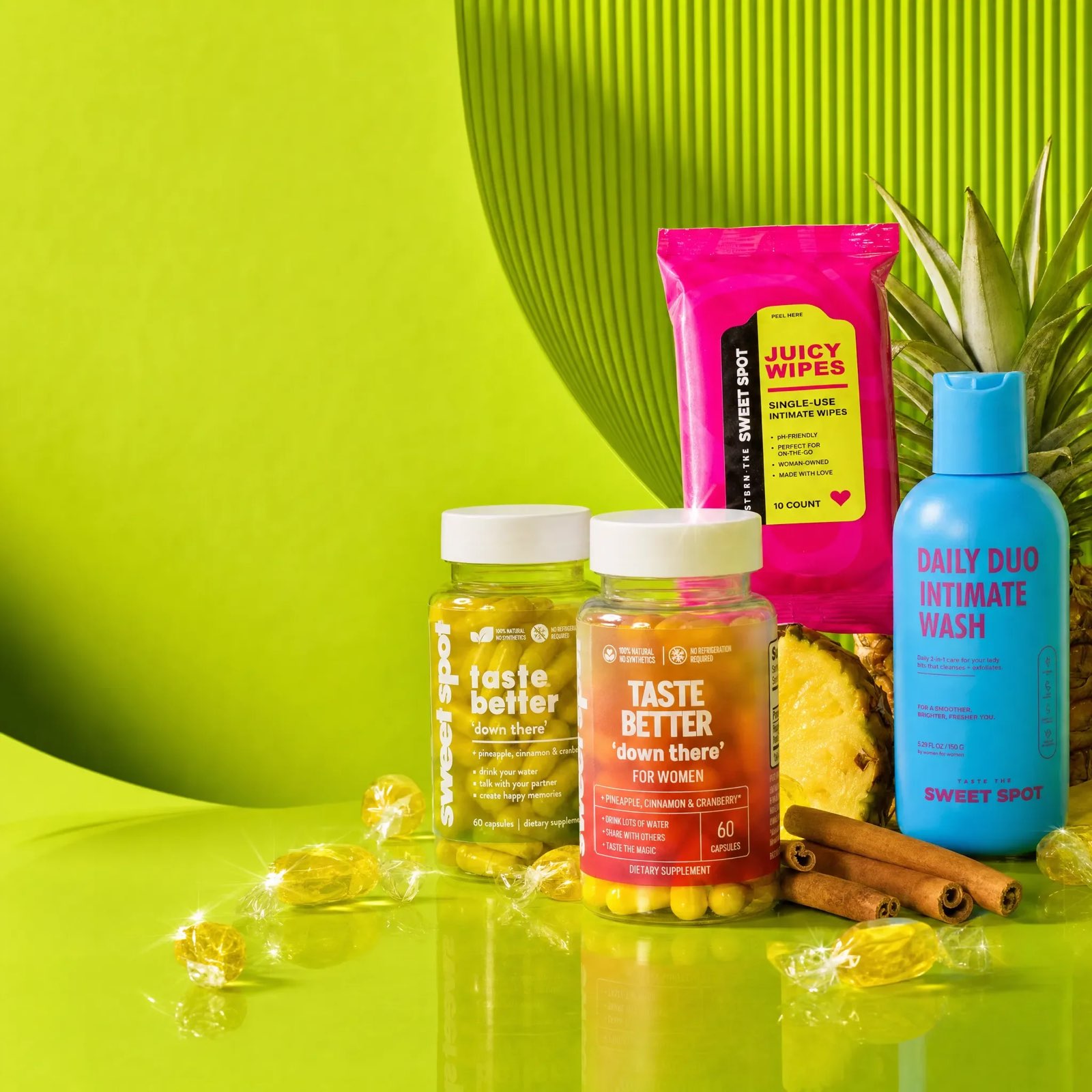 A green taste better down there bottle and an orange For Women bottle stand with pink Juicy Wipes, a blue Daily Duo wash, pineapple, and cinnamon on lime.