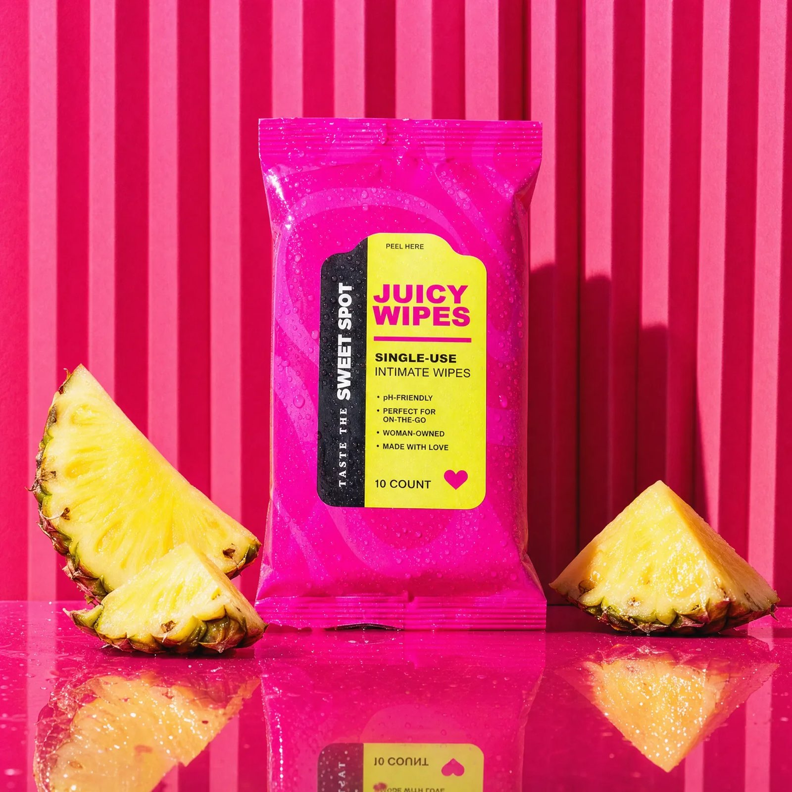 A pink Sweet Spot Juicy Wipes packet stands between fresh pineapple wedges on a glossy pink surface against matching vertical stripes.