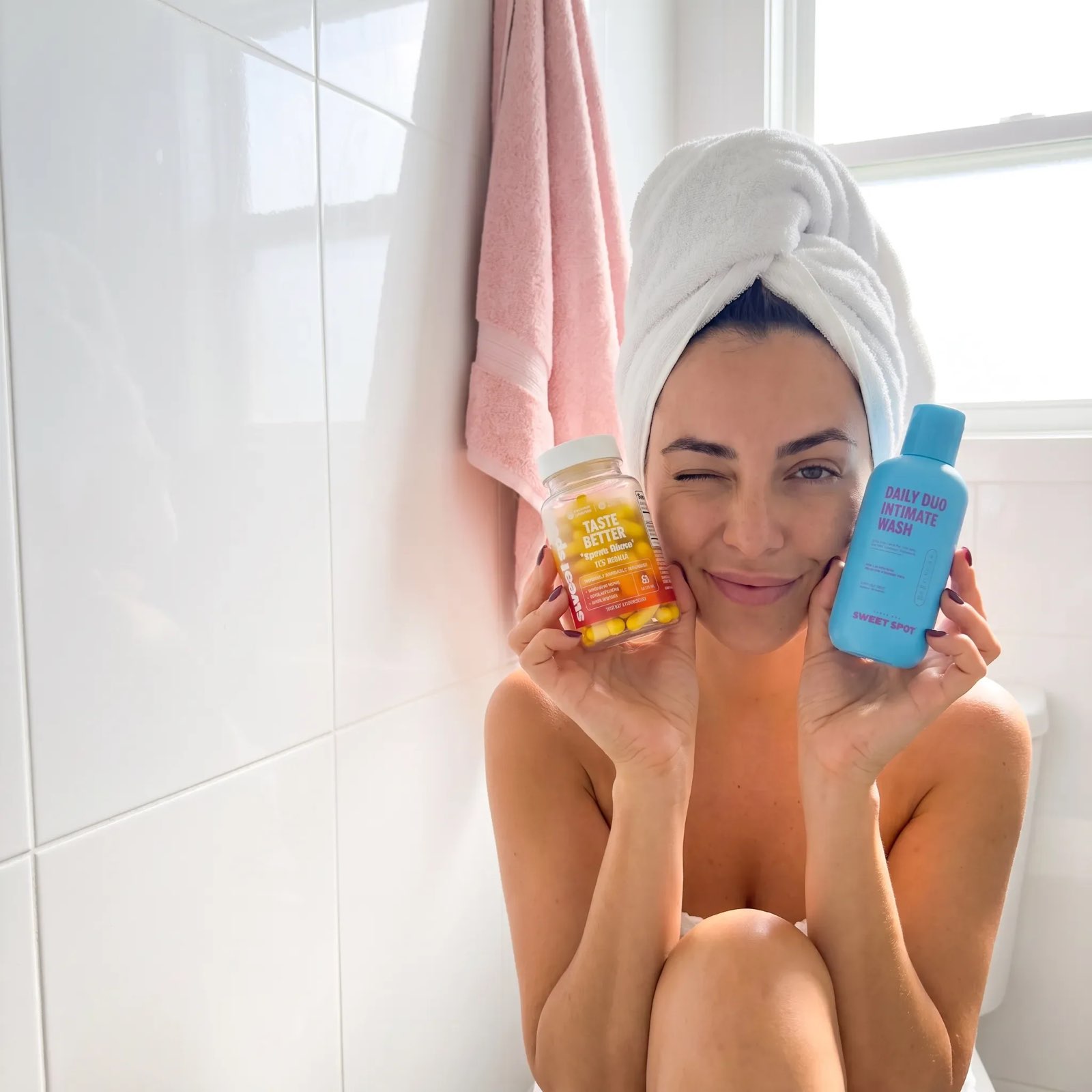 A seated woman in a white towel turban winks in a tiled bathroom while holding a Taste Better bottle and a blue Daily Duo wash.