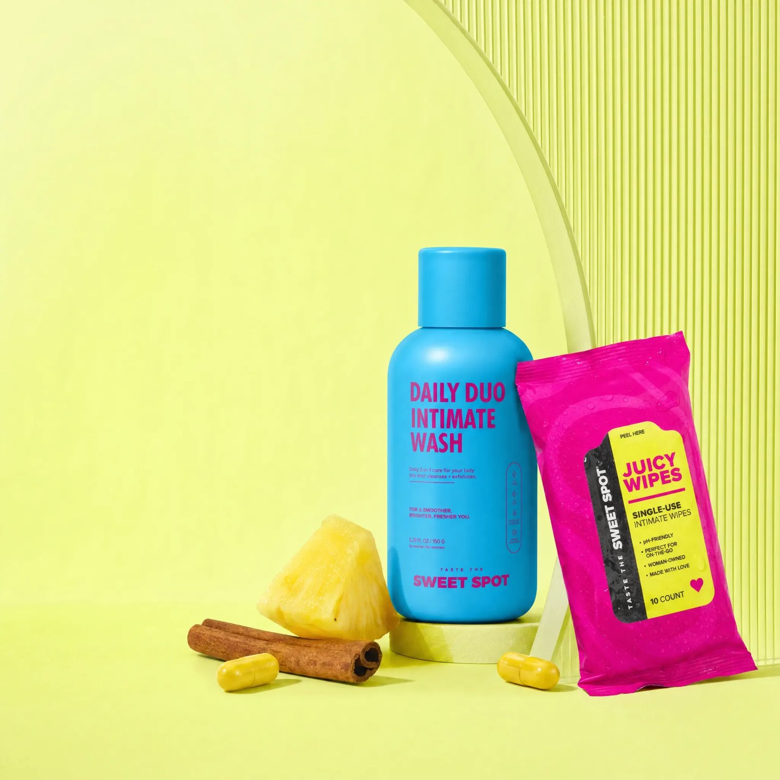 A blue Daily Duo Intimate Wash bottle stands beside a pink Juicy Wipes packet, a pineapple chunk, cinnamon sticks, and two yellow capsules on pale yellow.