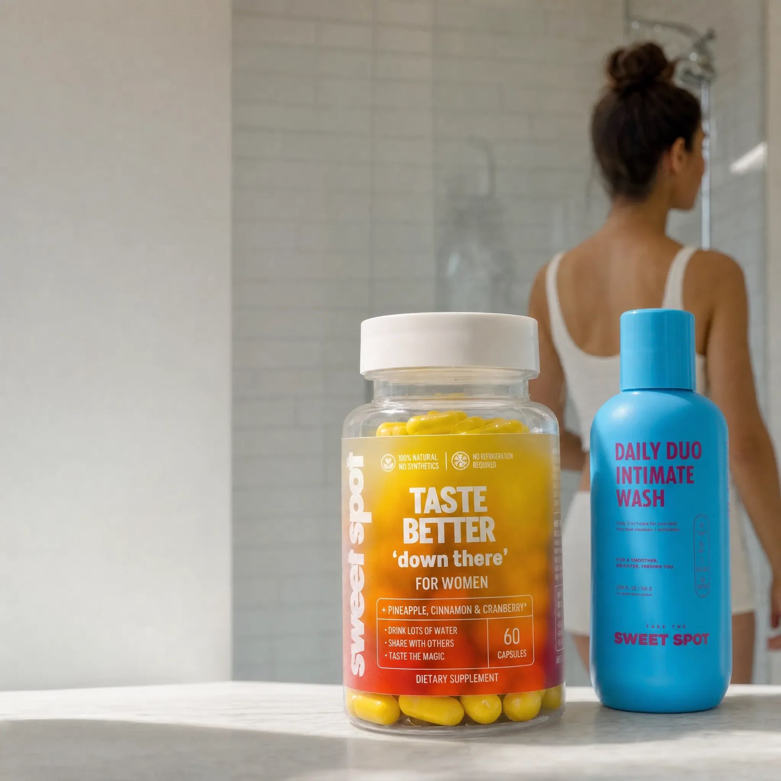 A Taste Better down there For Women bottle and a blue Daily Duo wash sit on marble while a woman in white faces a shower.