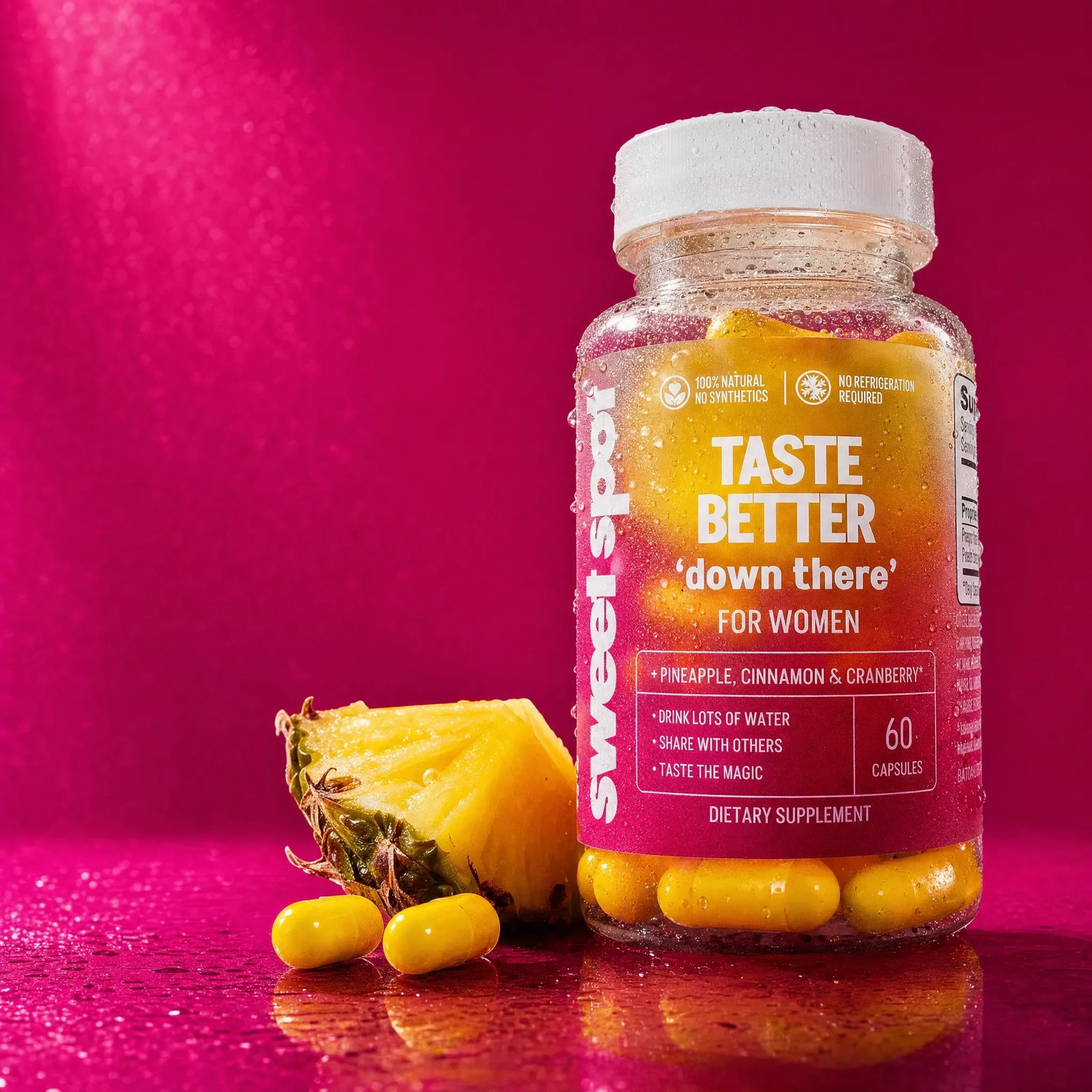 A dewy Sweet Spot Taste Better down there For Women bottle stands on wet magenta beside a pineapple wedge and two loose yellow capsules.