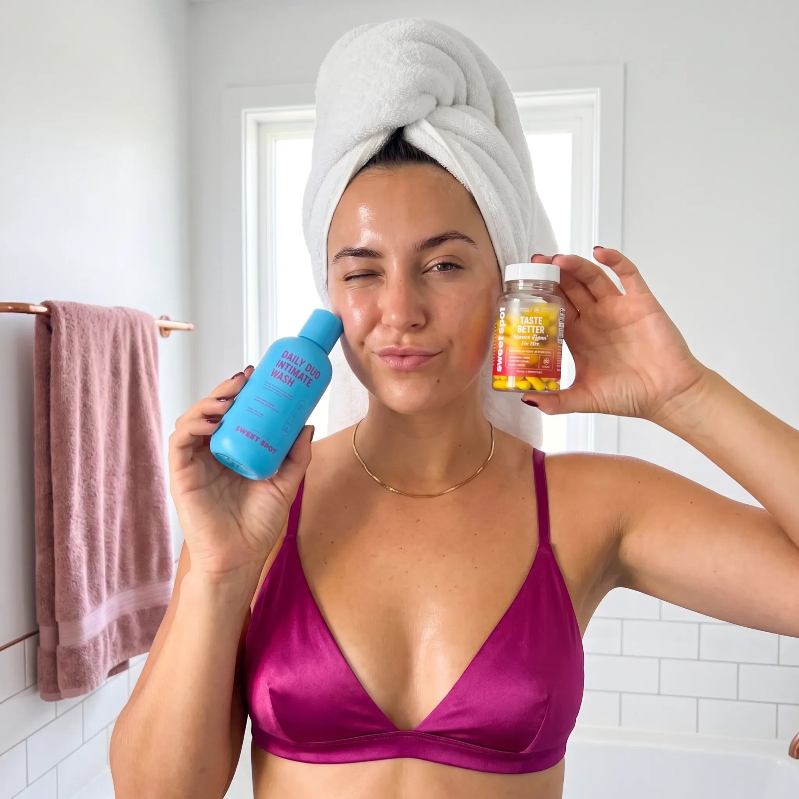 A woman with a towel on her head winks in a bathroom, holding blue Daily Duo Intimate Wash and a Taste Better down there for women bottle.