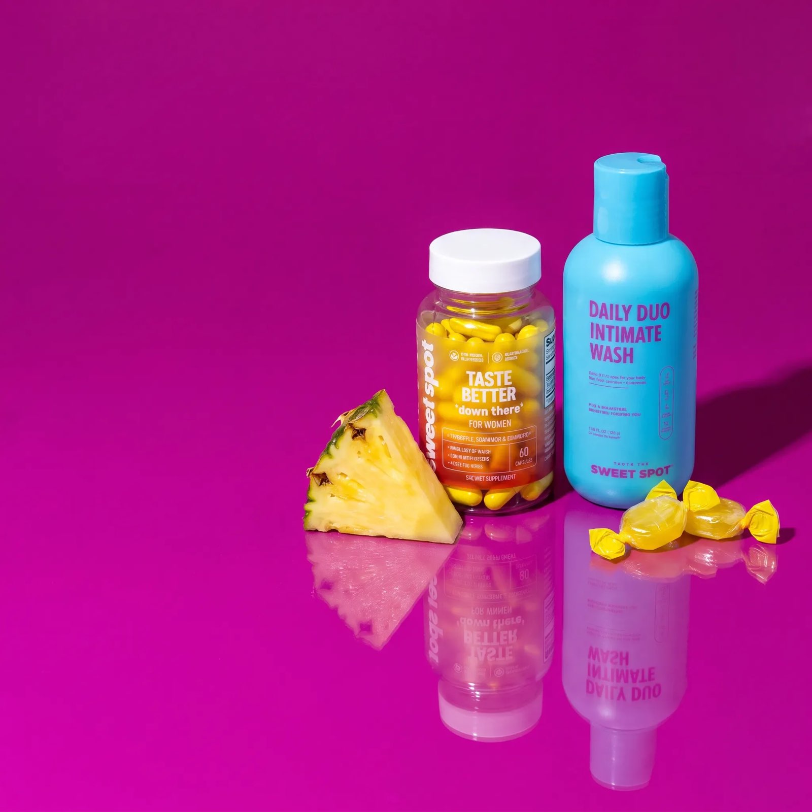 A pineapple wedge, a Taste Better down there for women bottle, and a blue Daily Duo Intimate Wash stand on a magenta surface with yellow candies.