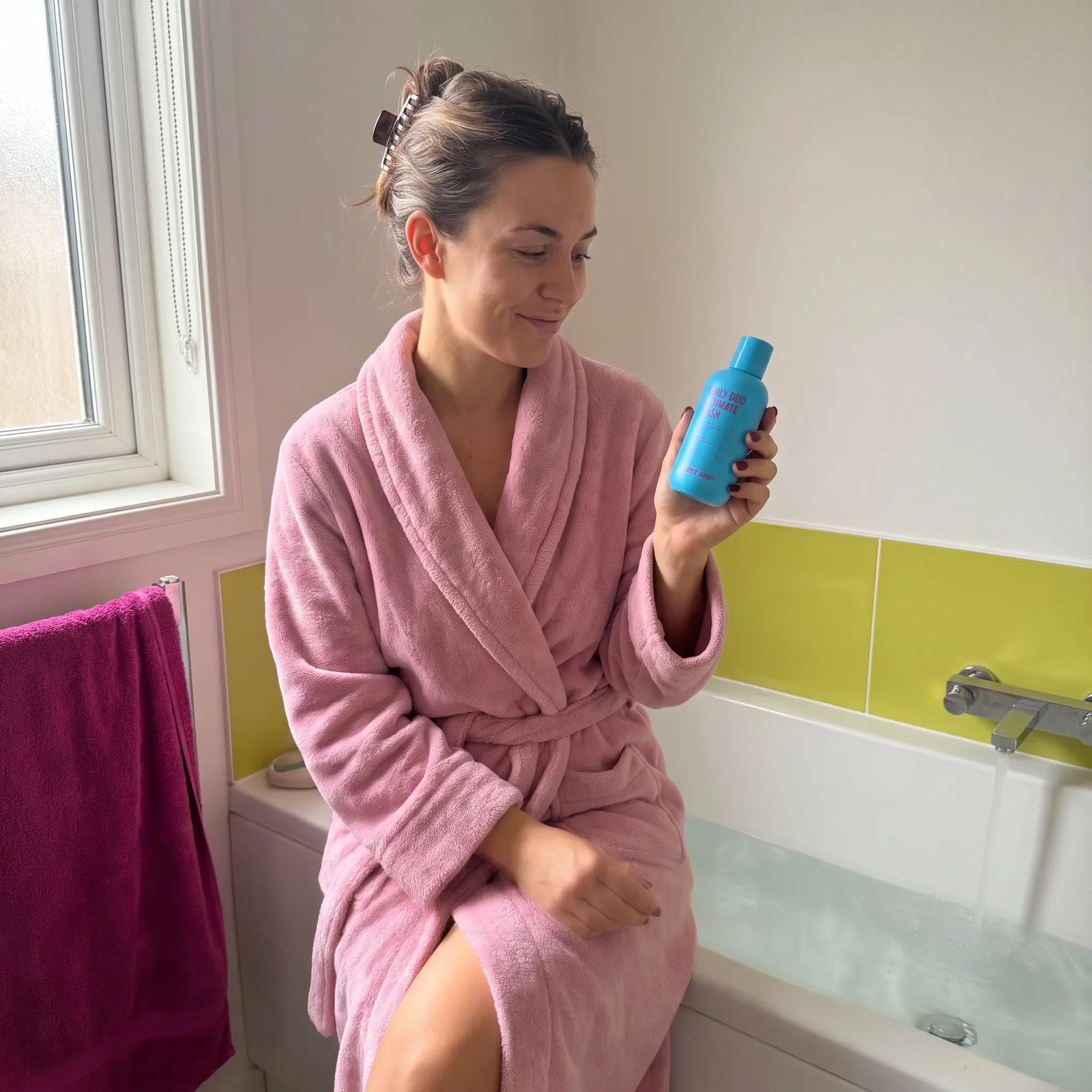 A woman in a pink robe sits on the edge of a filled tub, looking at a blue Daily Duo Intimate Wash as water runs.