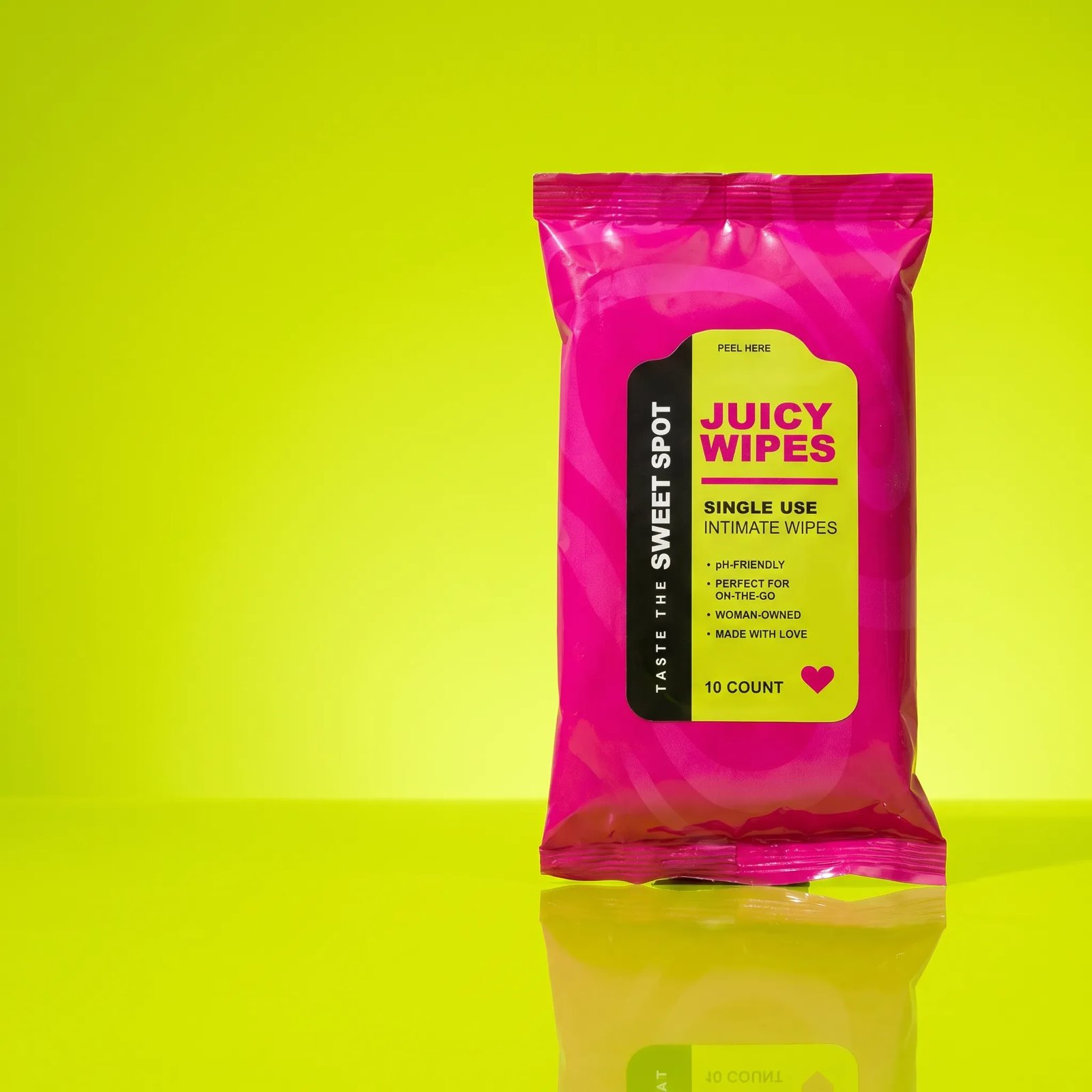 A pink Juicy Wipes packet labeled Taste the Sweet Spot stands alone on a glossy lime surface with its reflection below.