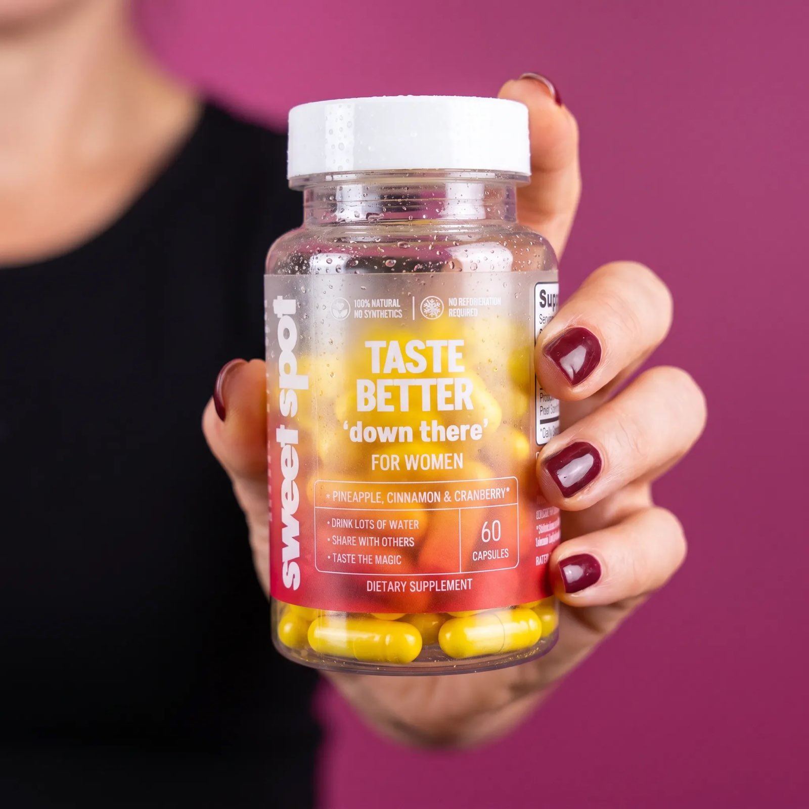A hand with dark red nails holds a droplet-covered Taste Better down there for women bottle filled with yellow capsules against a magenta backdrop.