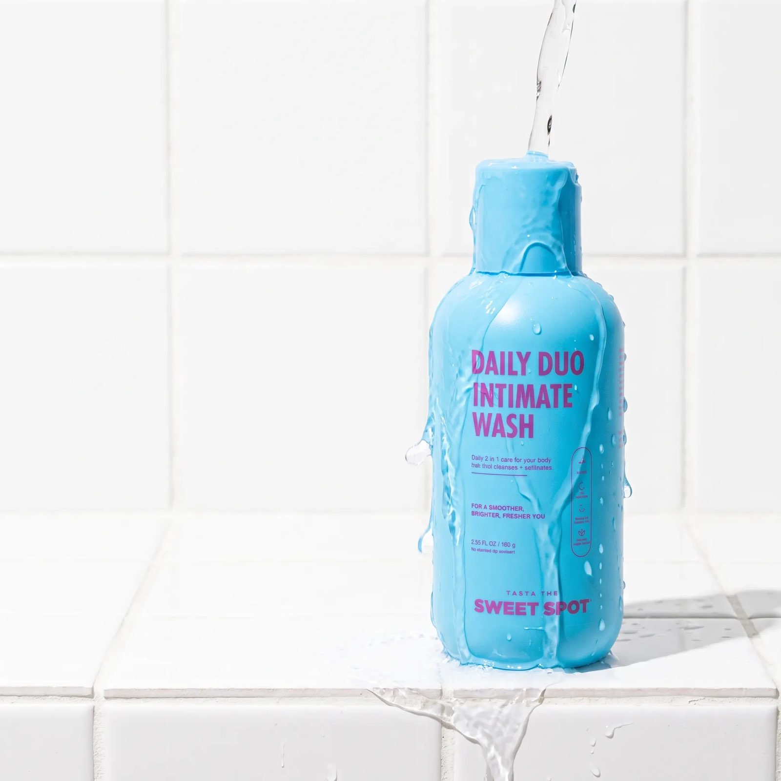 Water pours over a blue Daily Duo Intimate Wash bottle standing on white tile, with streams running down the label and off the ledge.