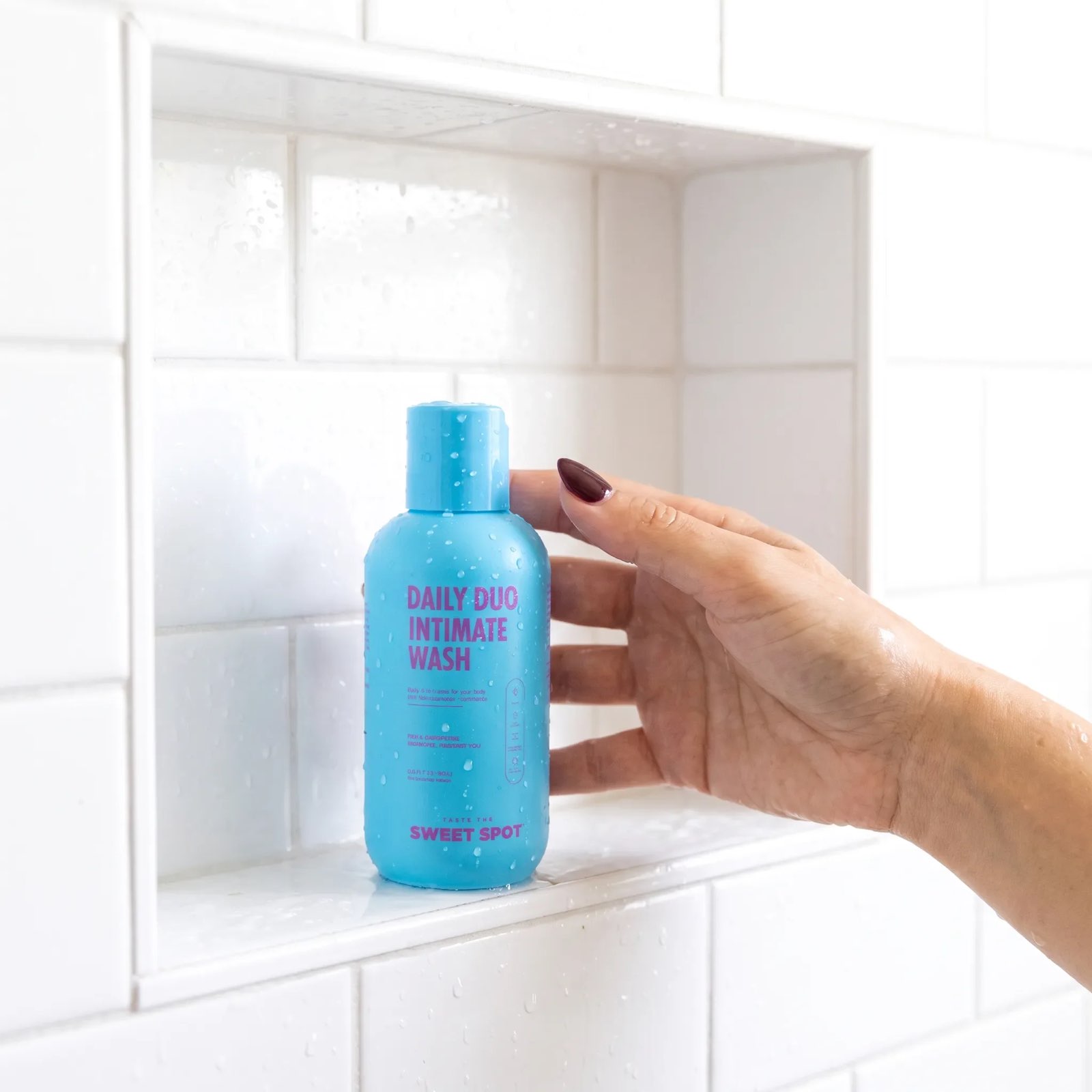 A wet hand with a dark red nail reaches toward a blue Daily Duo Intimate Wash bottle sitting in a white tiled shower niche.