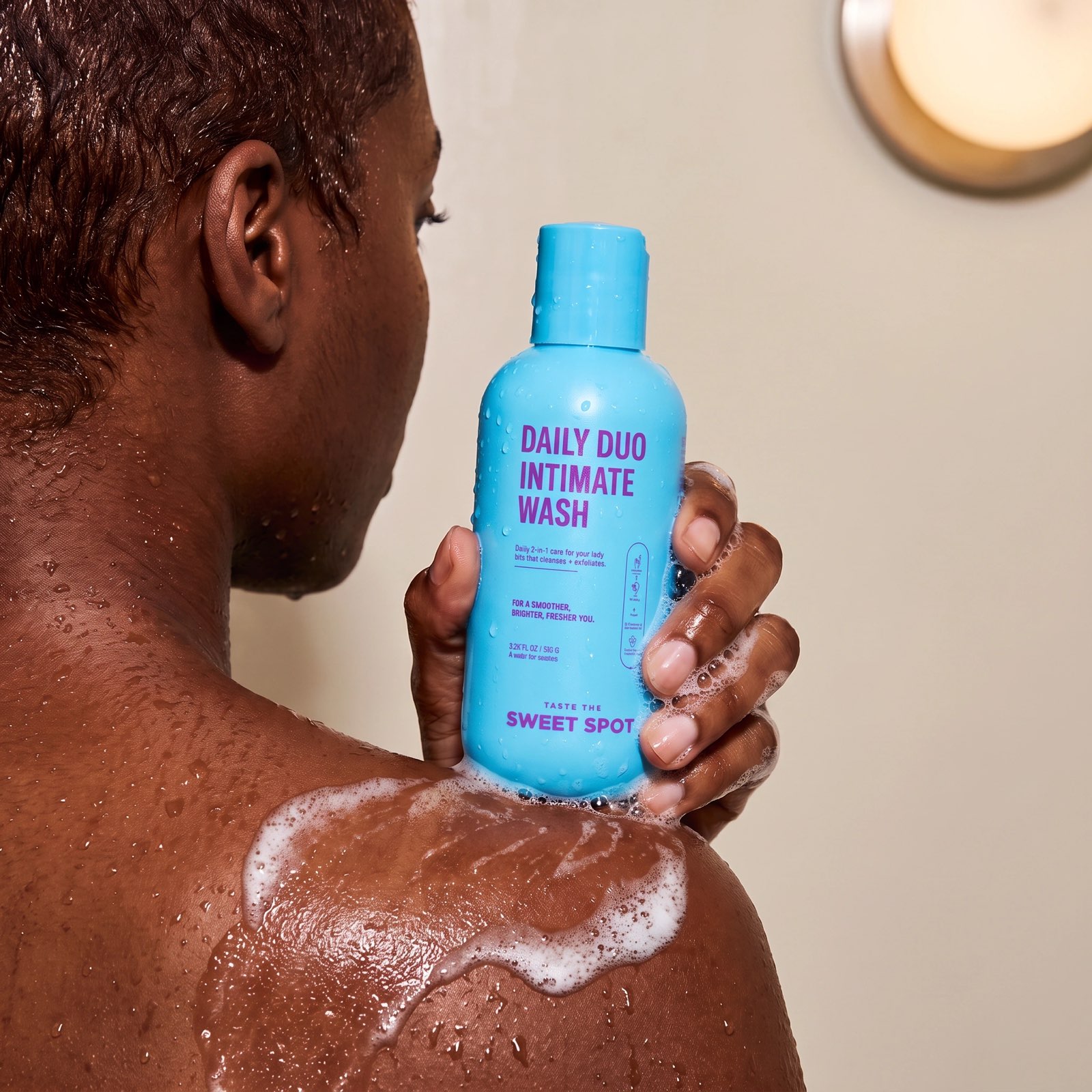From behind, a person with wet skin and soap on one shoulder holds a blue Daily Duo Intimate Wash bottle in a shower.
