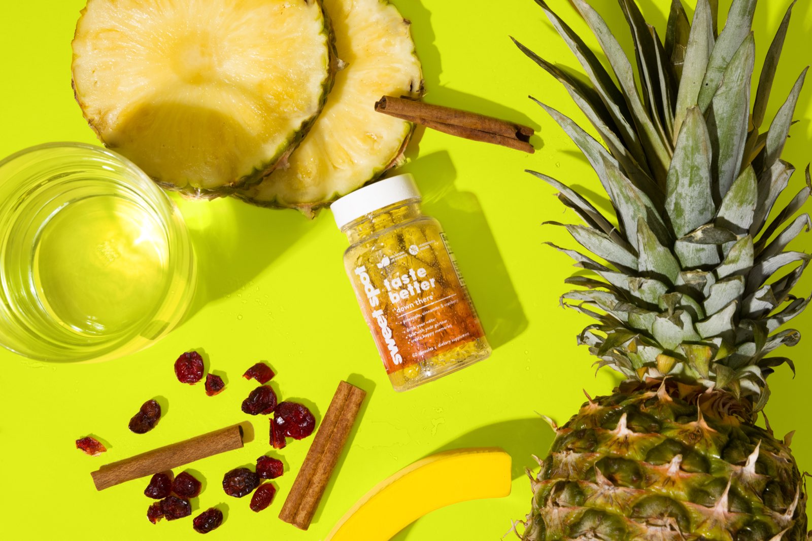 On a lime background, a Sweet Spot bottle labeled taste better down there sits with pineapple rings, a whole pineapple, cranberries, cinnamon, and a glass.