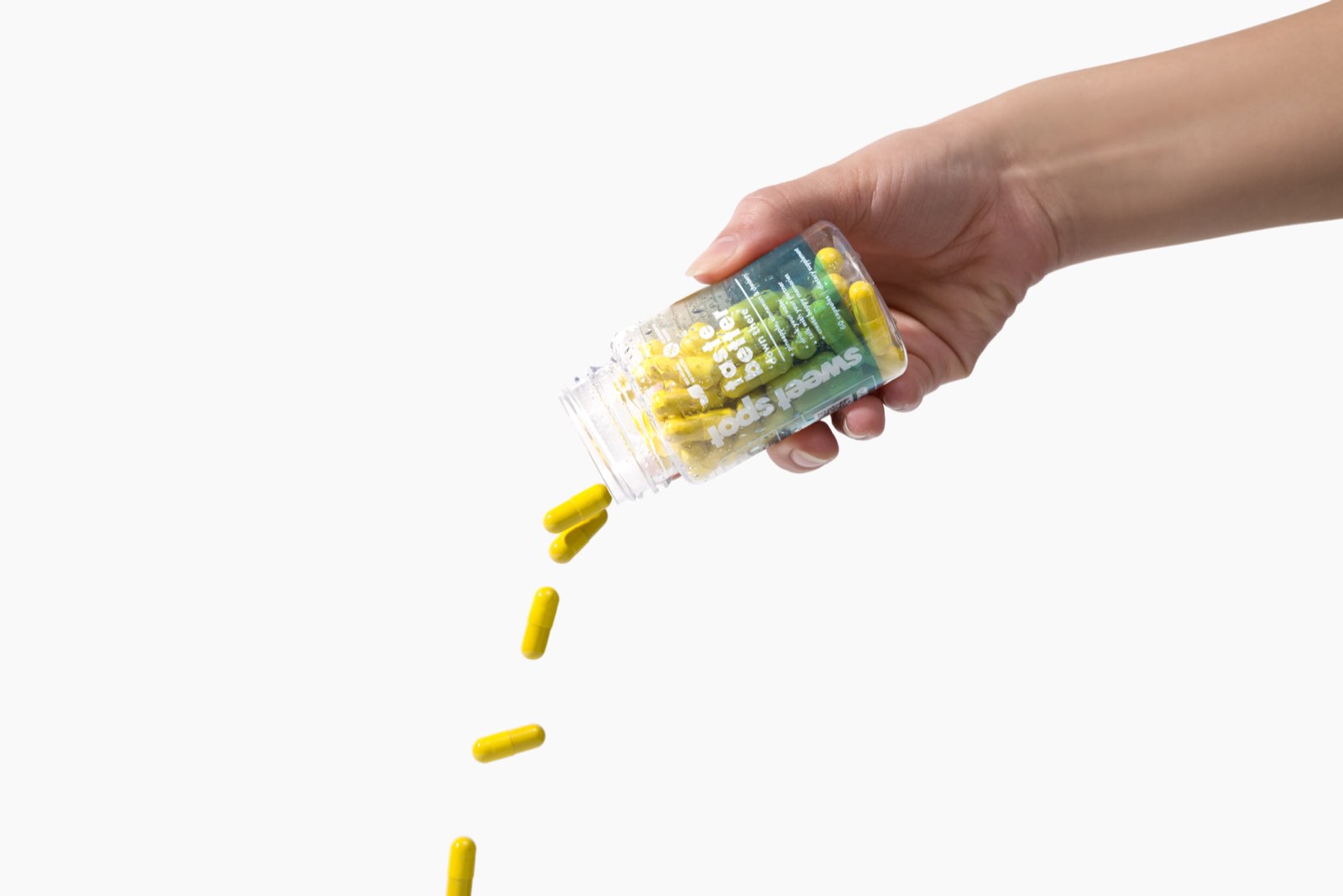 A hand pours yellow capsules from a green-labeled Sweet Spot bottle reading taste better down there, on a white background.