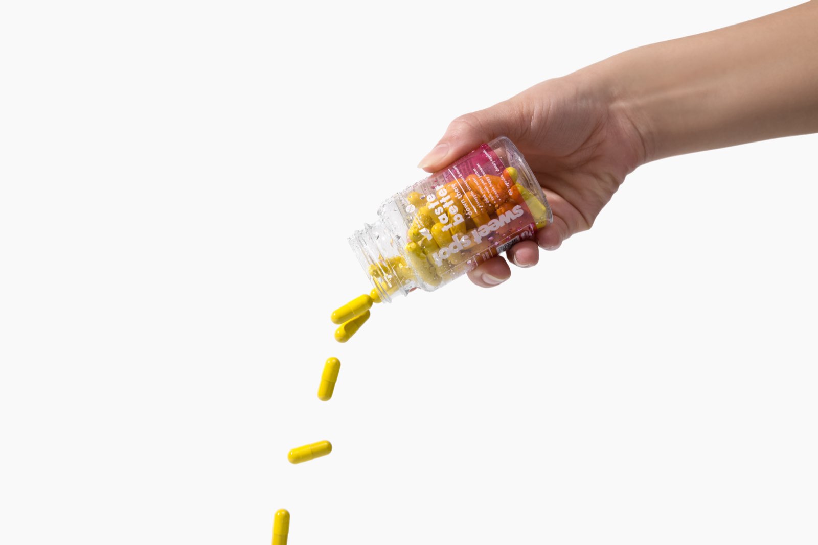 A hand pours yellow capsules from a pink-labeled Sweet Spot bottle reading taste better, with orange capsules still inside against a white background.