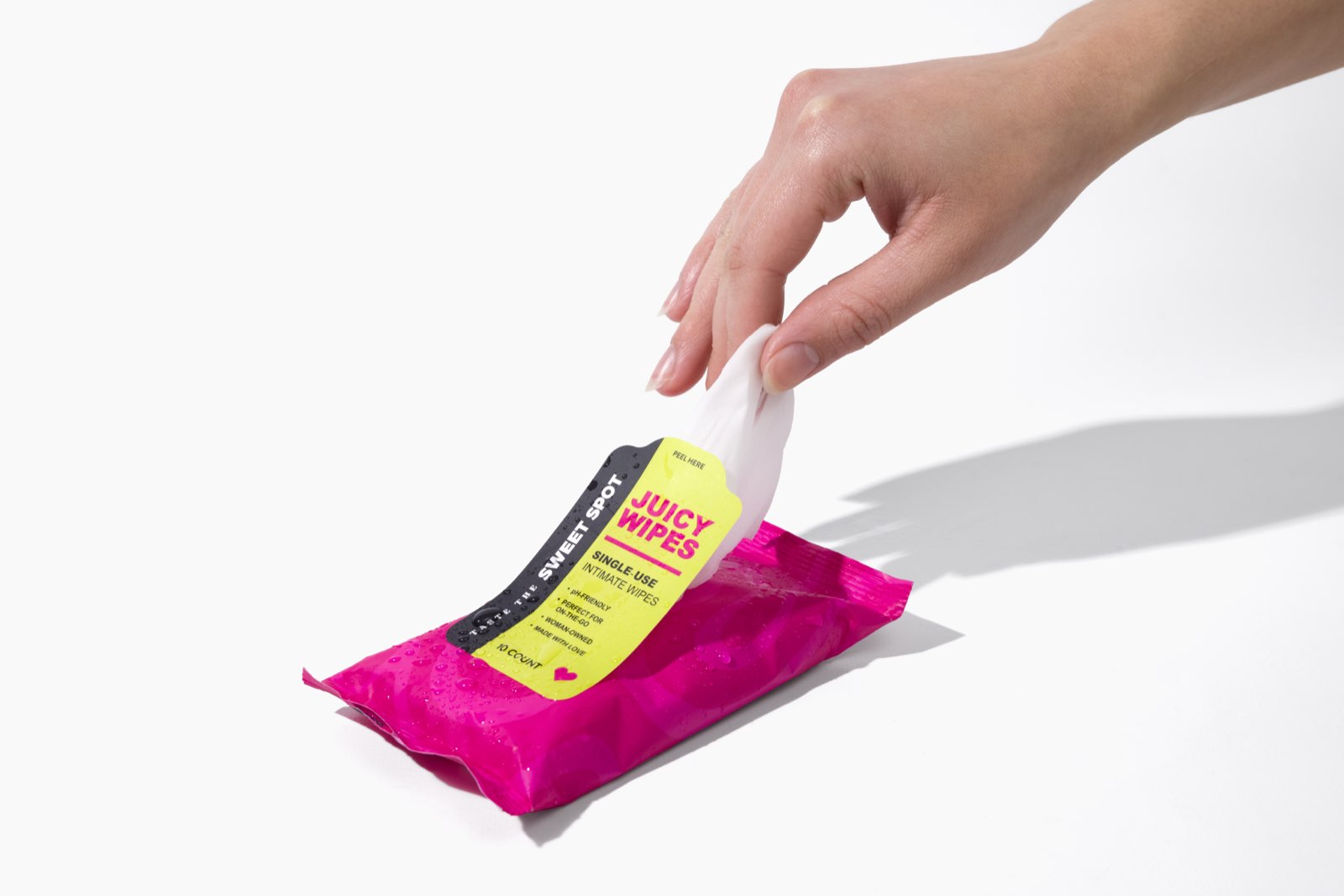 A hand pulls a white wipe from a pink Juicy Wipes packet labeled single-use intimate wipes and Taste the Sweet Spot on a white surface.