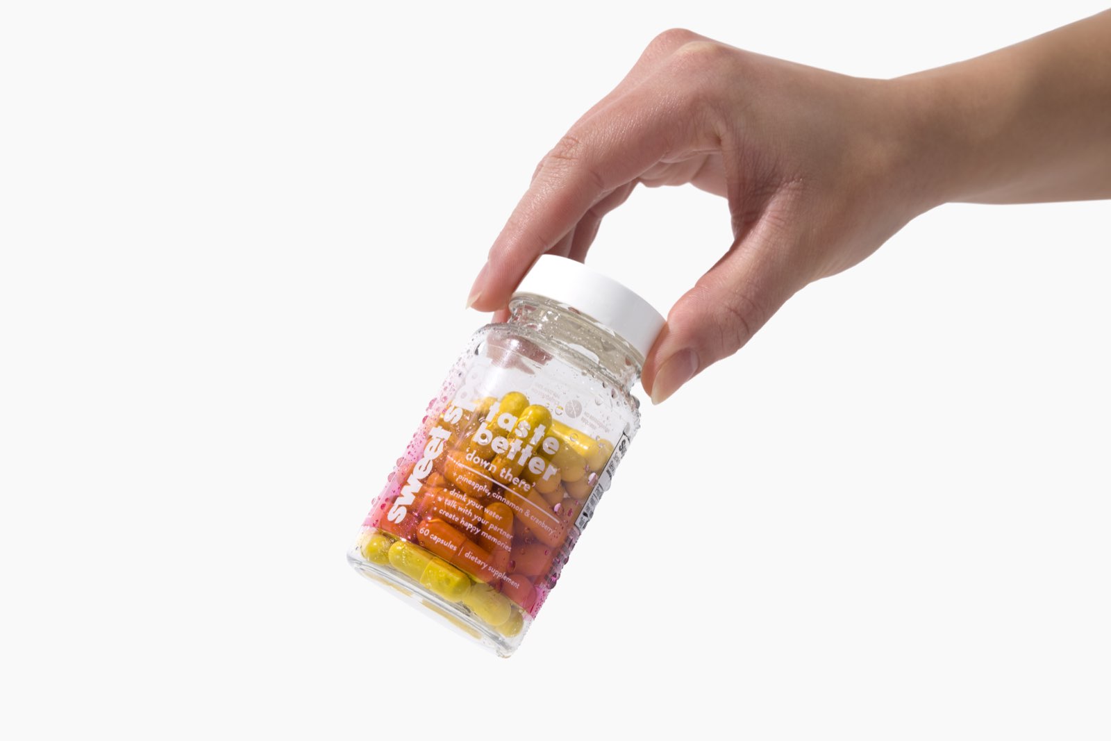 A hand holds a wet pink-labeled Sweet Spot bottle reading taste better down there, yellow and orange capsules showing through the clear plastic.