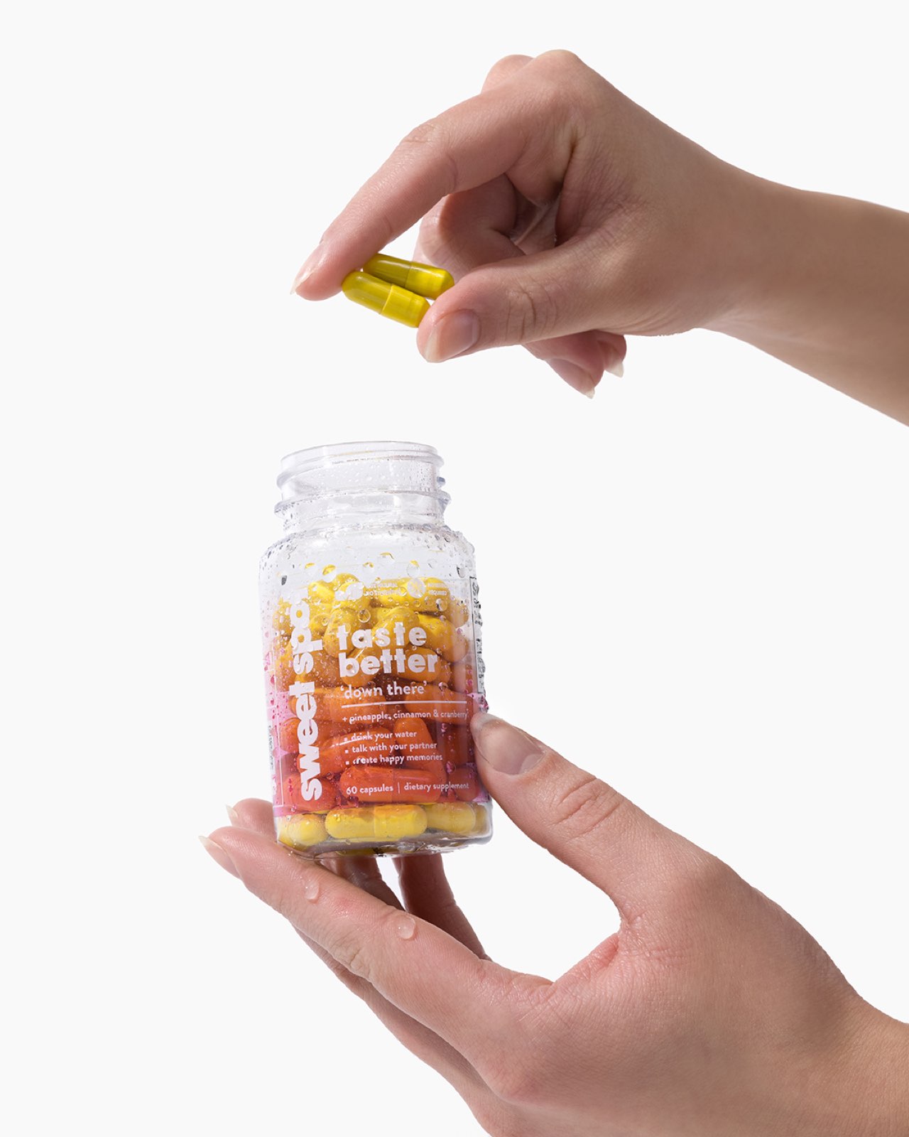 Two hands cradle an open pink-labeled Sweet Spot bottle reading taste better down there while fingers pinch two yellow capsules just above the neck.