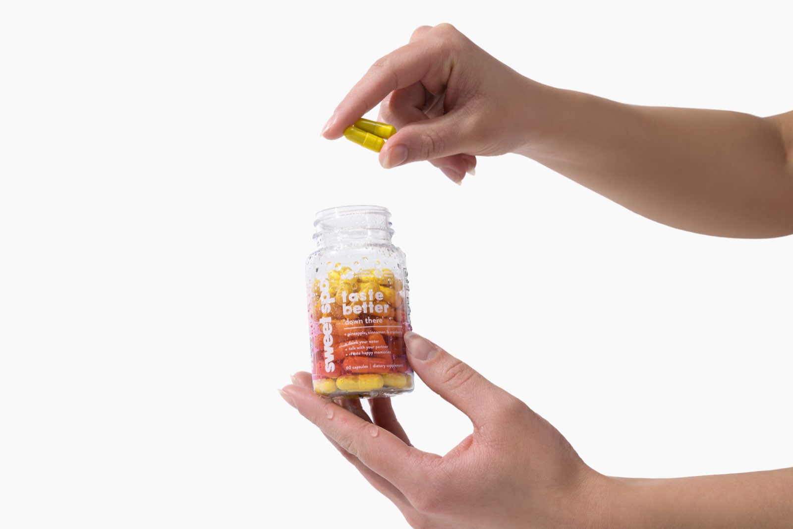 An open palm supports a wet pink-labeled Sweet Spot bottle reading taste better down there while two yellow capsules are held above the mouth.