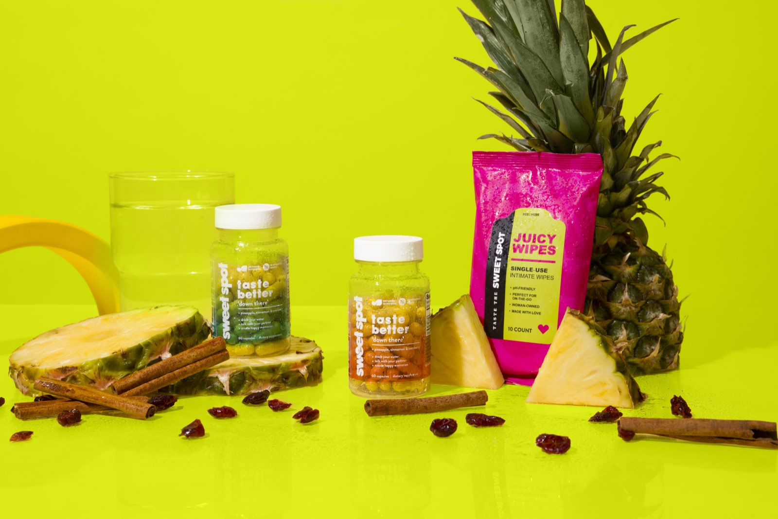 A green Sweet Spot bottle and an orange taste better down there bottle stand among pineapple, cinnamon, cranberries, a water glass, and a pink Juicy Wipes packet.