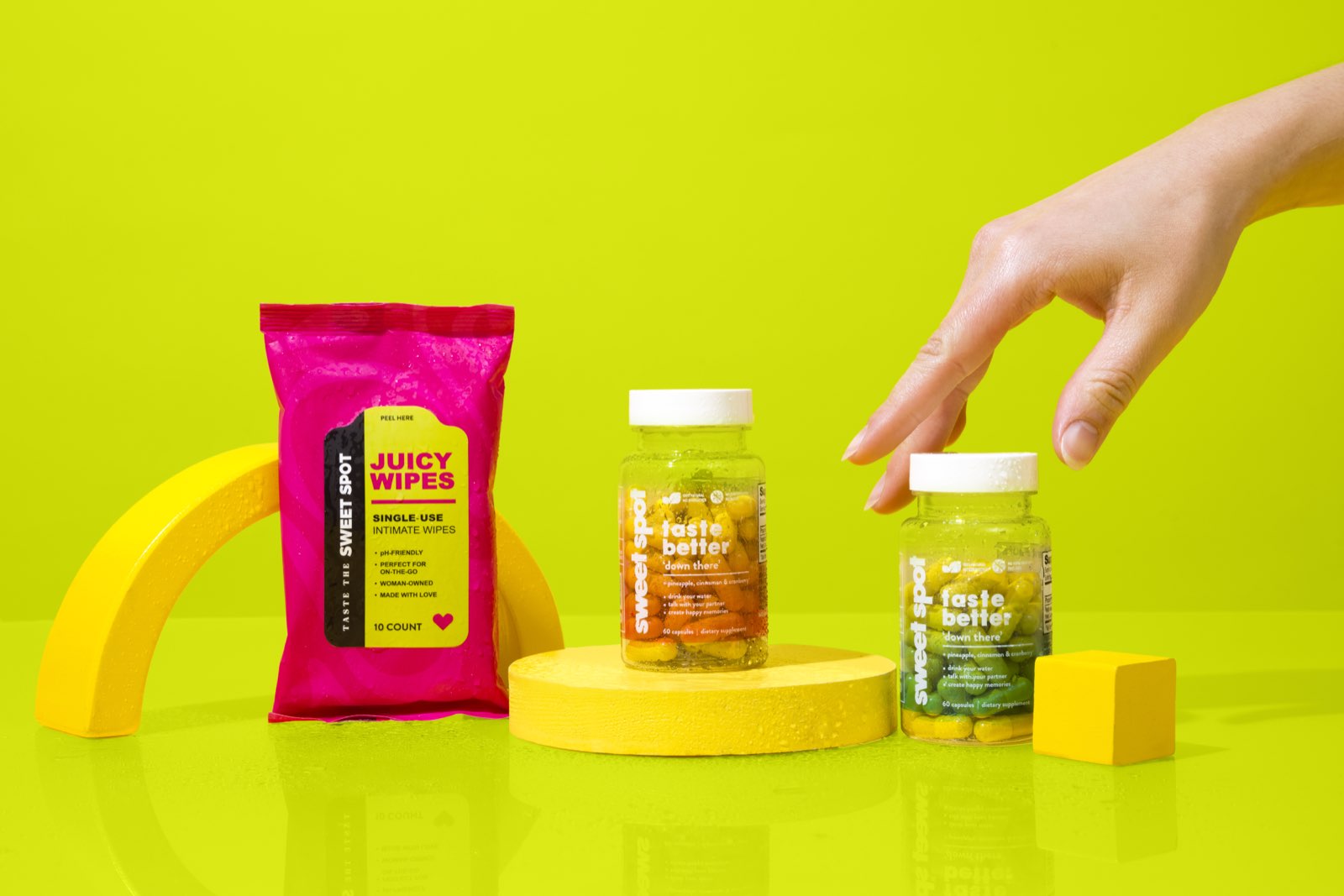 A hand reaches toward the green Sweet Spot bottle beside an orange taste better down there bottle on a yellow podium and a pink Juicy Wipes packet.