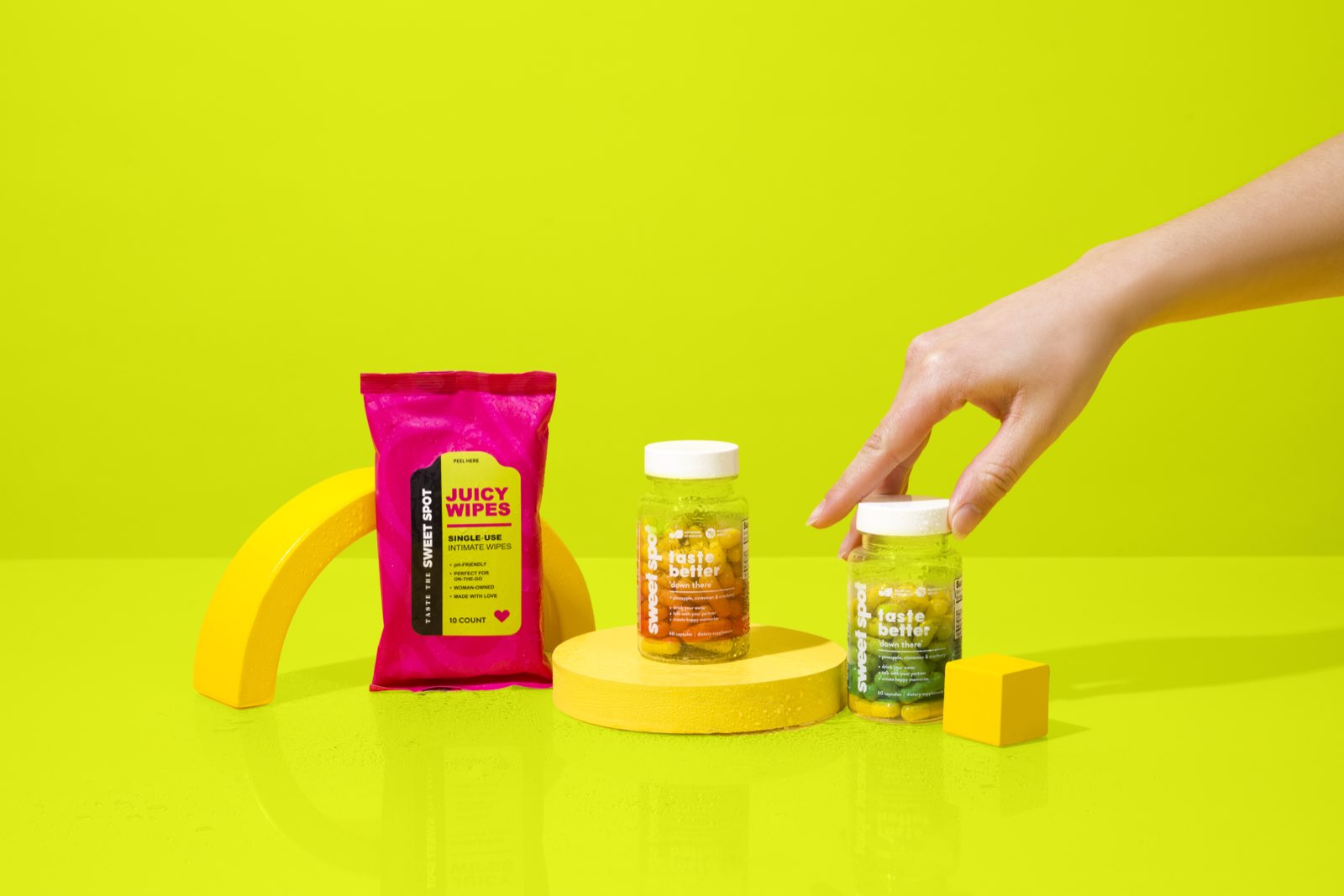 A hand grasps the cap of the green Sweet Spot bottle next to an orange taste better down there bottle, a pink Juicy Wipes packet, and yellow props.
