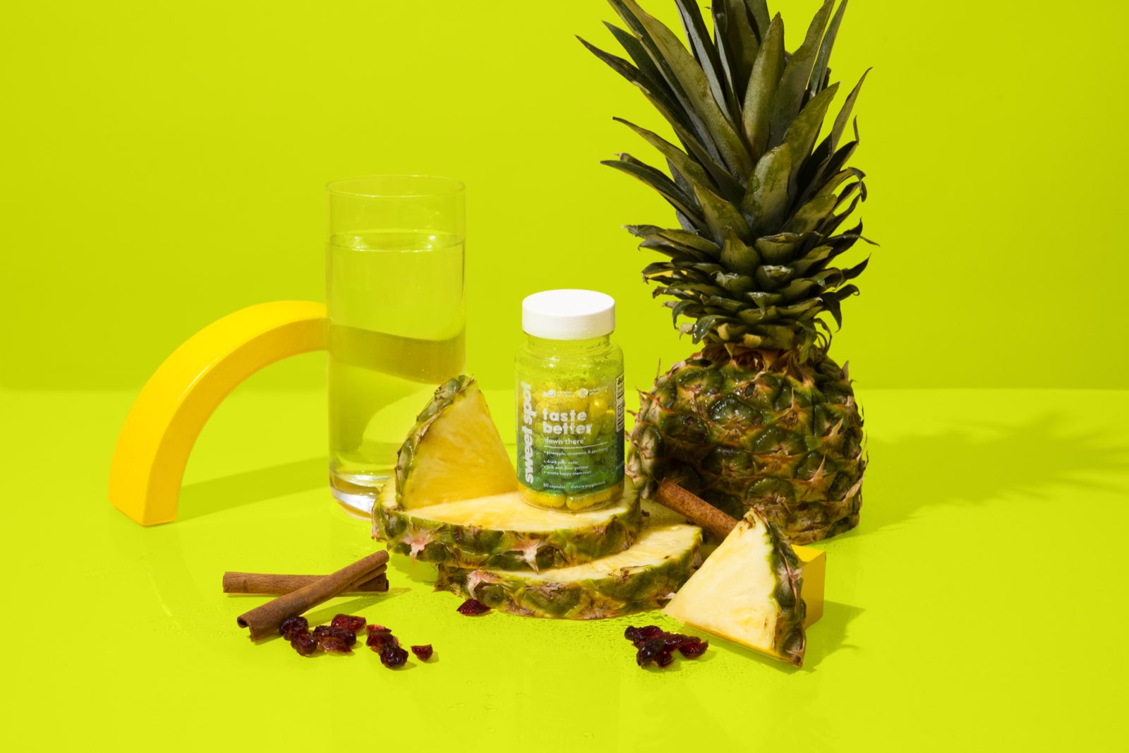 A green Sweet Spot taste better down there bottle sits among pineapple slices, a whole pineapple, cinnamon sticks, cranberries, and a tall glass of water.