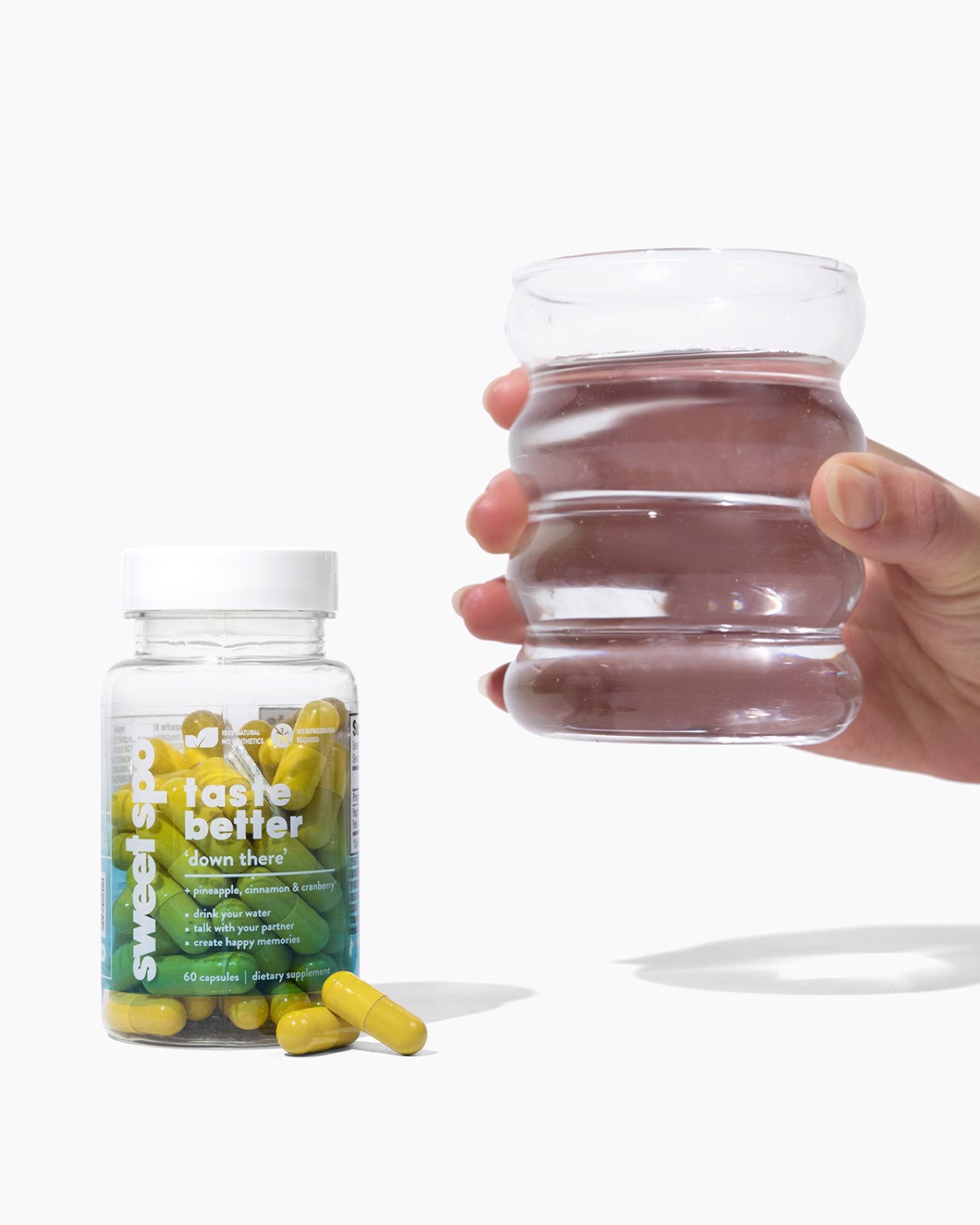 A hand holds a ribbed glass of water beside a green Sweet Spot taste better down there bottle, with yellow capsules lying in front on white.