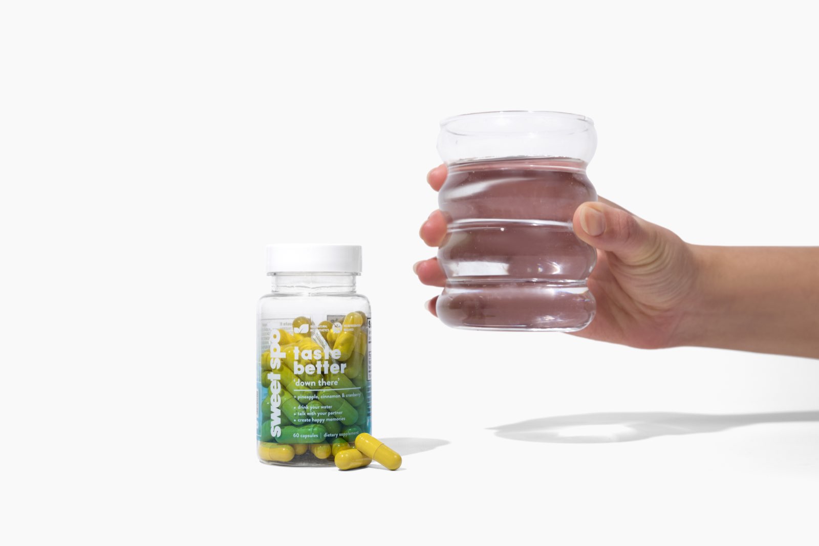 On a white surface, a green Sweet Spot taste better down there bottle and two yellow capsules sit beside a hand holding a ribbed glass of water.