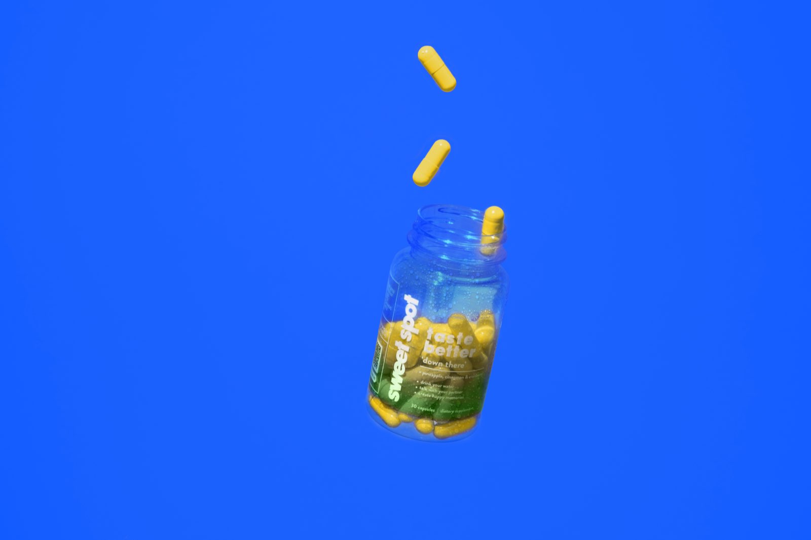 An open green Sweet Spot taste better down there bottle tilts on a blue background while yellow capsules float above its mouth.