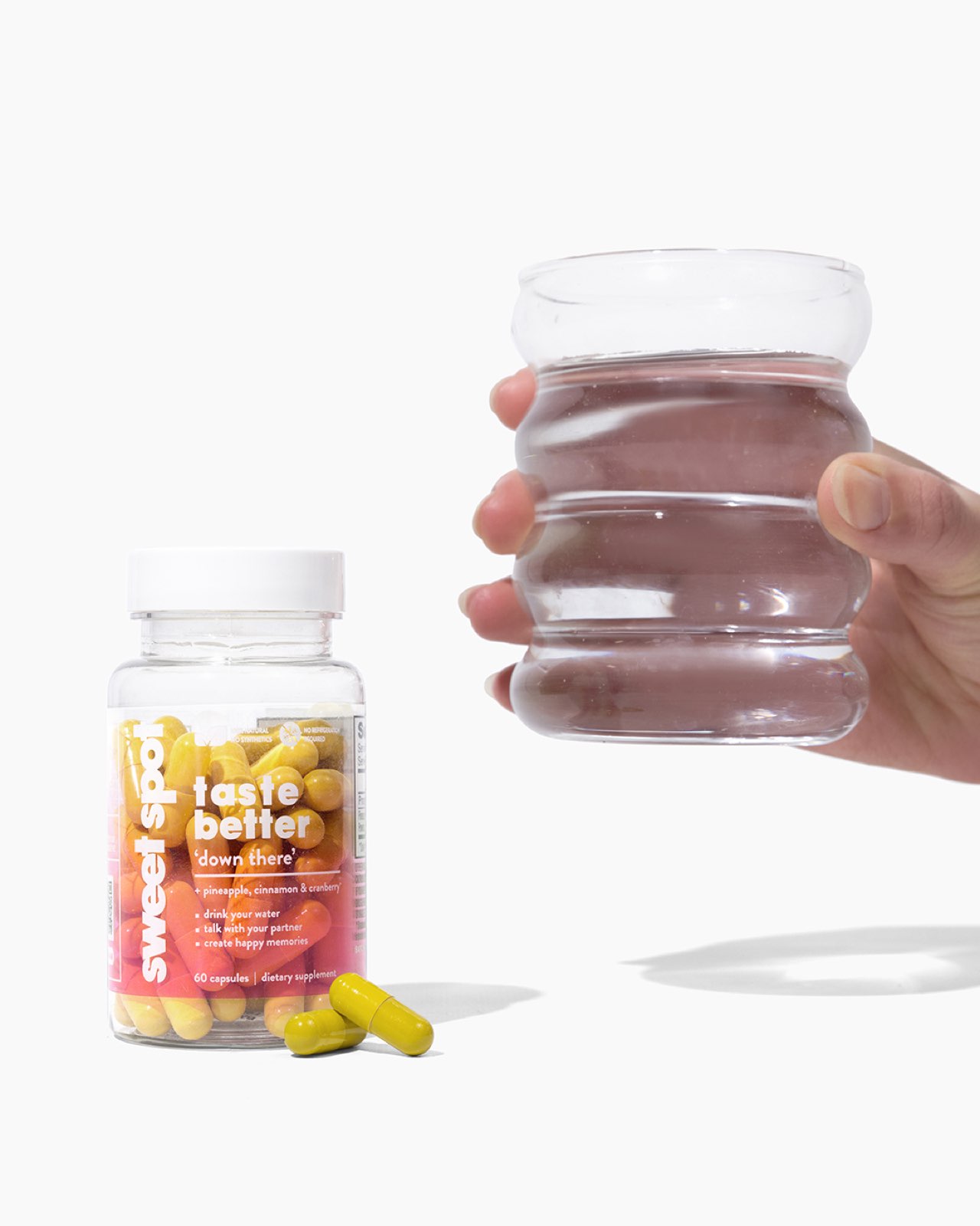 A hand holds a ribbed glass of water beside an orange Sweet Spot taste better down there bottle, with yellow capsules in front on white.