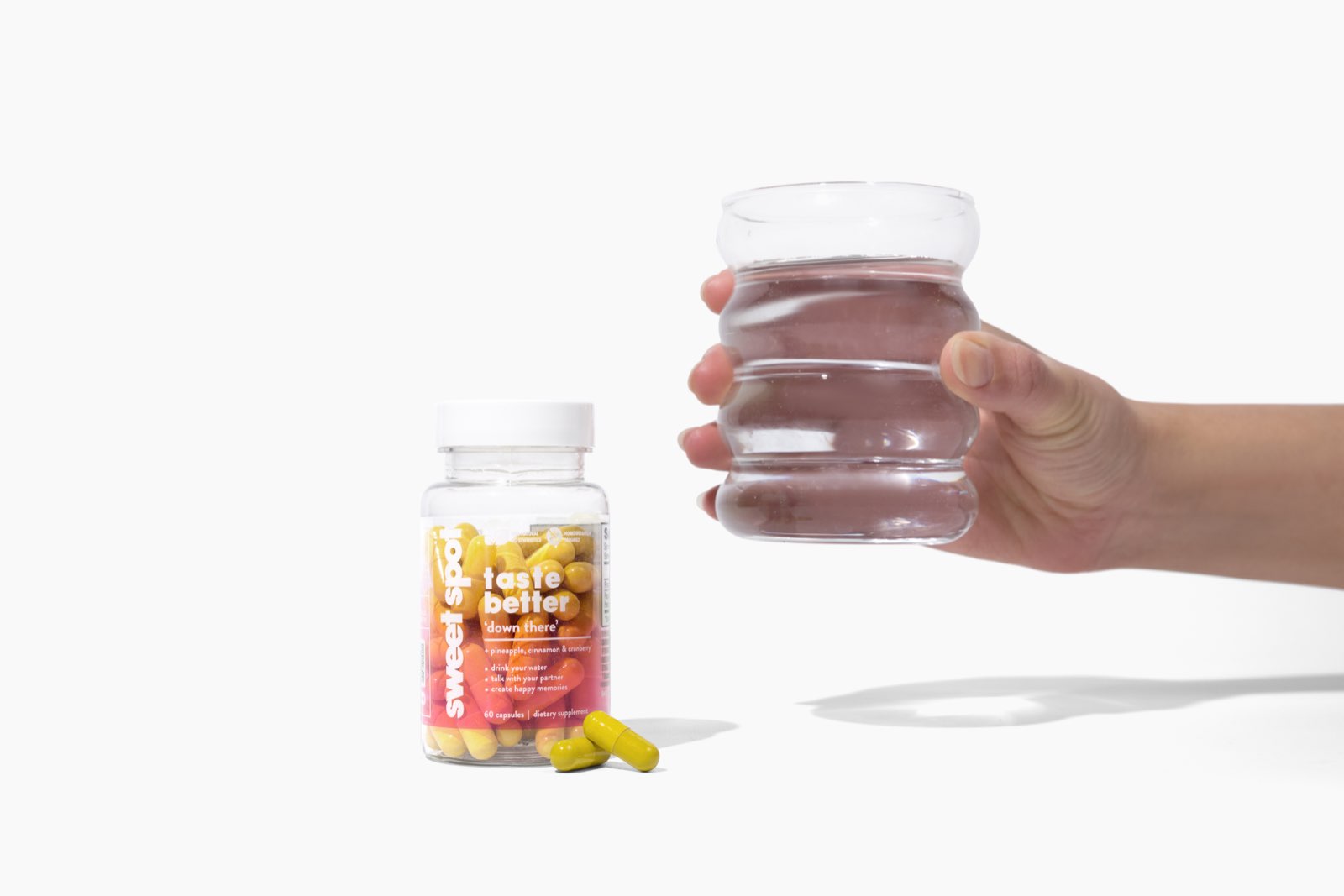 On white, an orange Sweet Spot taste better down there bottle and two yellow capsules sit beside a hand holding a ribbed glass of water.