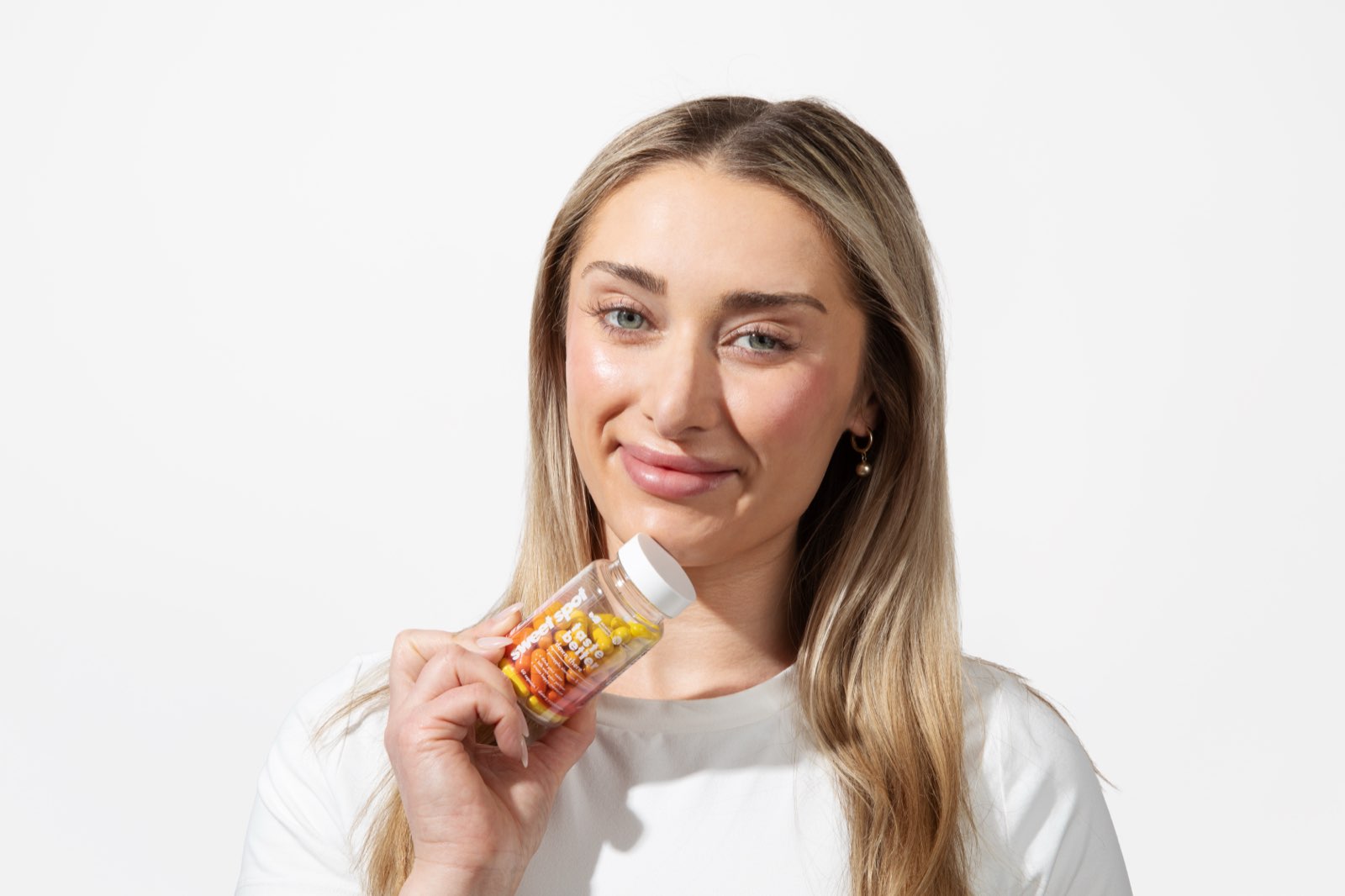 A blonde woman in a white shirt smiles at the camera while holding an orange Sweet Spot taste better down there bottle near her chin.