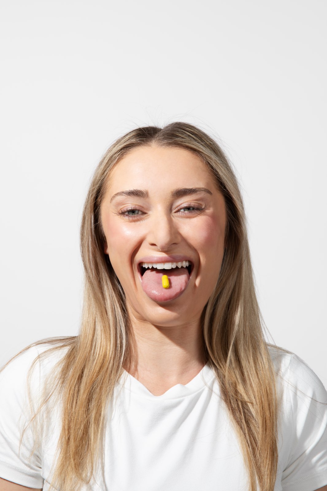 A blonde woman in a white shirt faces the camera with her tongue out and a yellow capsule resting on it.